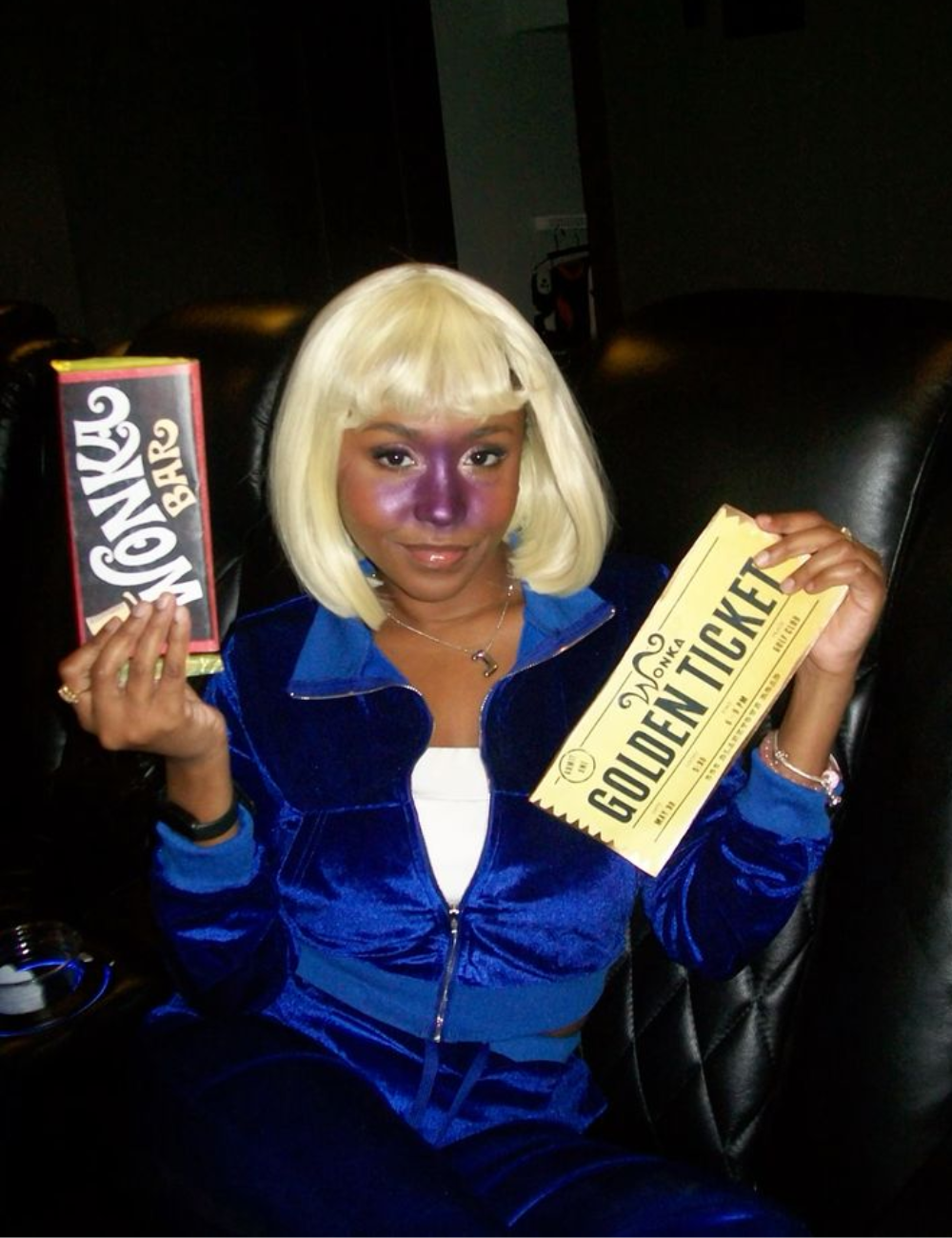 A imagem apresenta uma jovem com cabelo loiro platinado e maquiagem que destaca um tom roxo no rosto, remetendo a uma fantasia criativa. Ela veste um conjunto azul de veludo, confortável e estiloso, segurando uma barra de chocolate