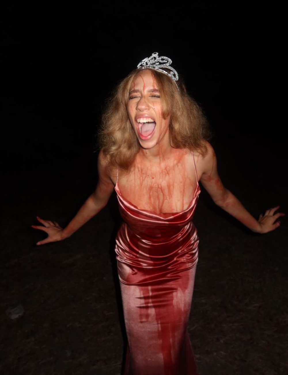 A imagem apresenta uma mulher com um vestido longo e justo, em tom de vermelho, que simula estar ensanguentado, ideal para fantasias de Halloween simples. Ela usa uma tiara brilhante e tem cabelos soltos e despenteados.