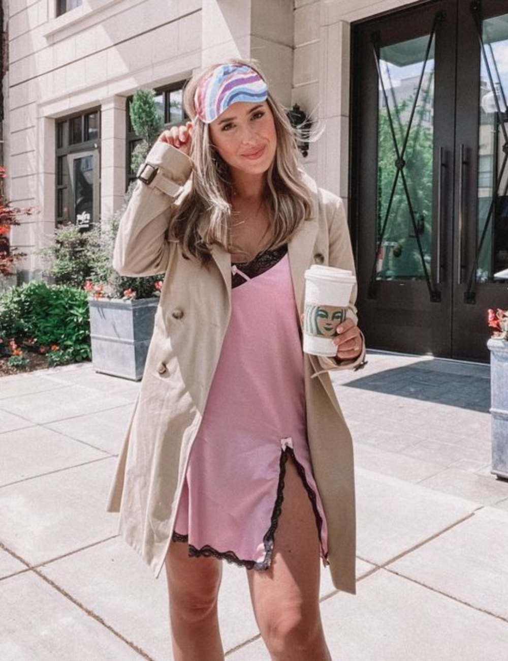 A imagem apresenta uma mulher usando uma fantasia de Halloween simples e criativa. Ela veste um vestido rosa com detalhes em renda preta, complementado por um trench coat bege. Um acessório divertido, uma faixa colorida para os cabelos.