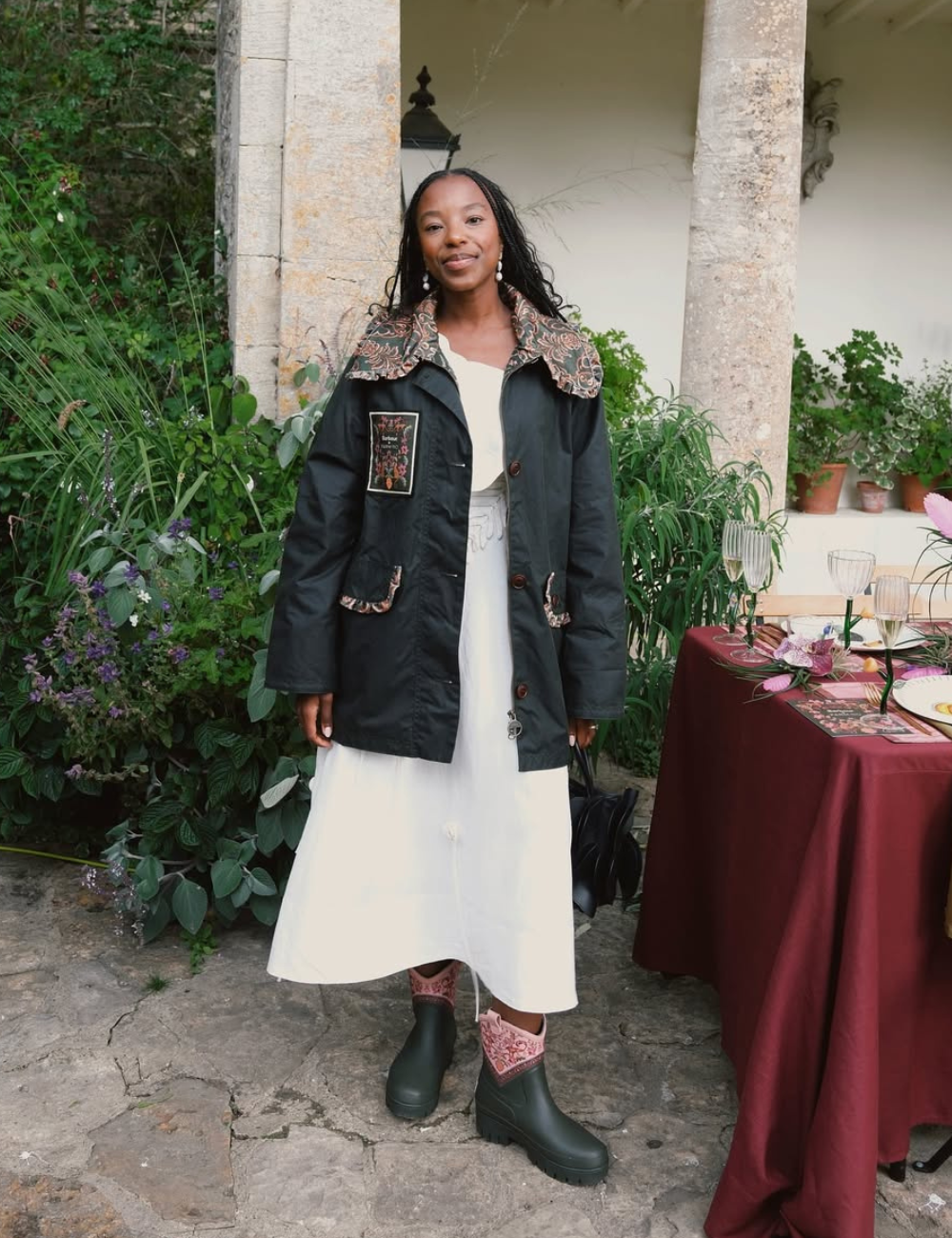 Look perfeito para dias chuvosos, apresentando uma combinação de estilo e funcionalidade. A modelo usa um casaco preto com detalhes bordados e um vestido branco fluido, ideal para camadas.