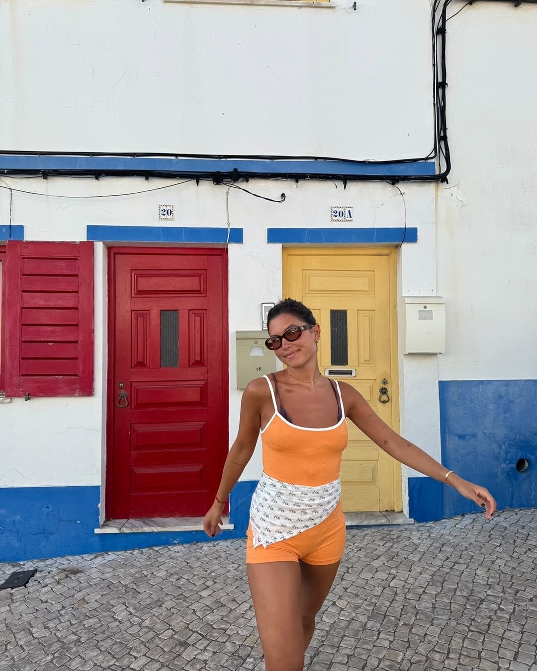 A imagem apresenta uma mulher usando um macacão estiloso em tons de laranja e branco, com detalhes em estampa na parte inferior. Ela está em um ambiente colorido, com portas vermelha e amarela ao fundo, que realçam a vivacidade do look. Óculos de sol sofi