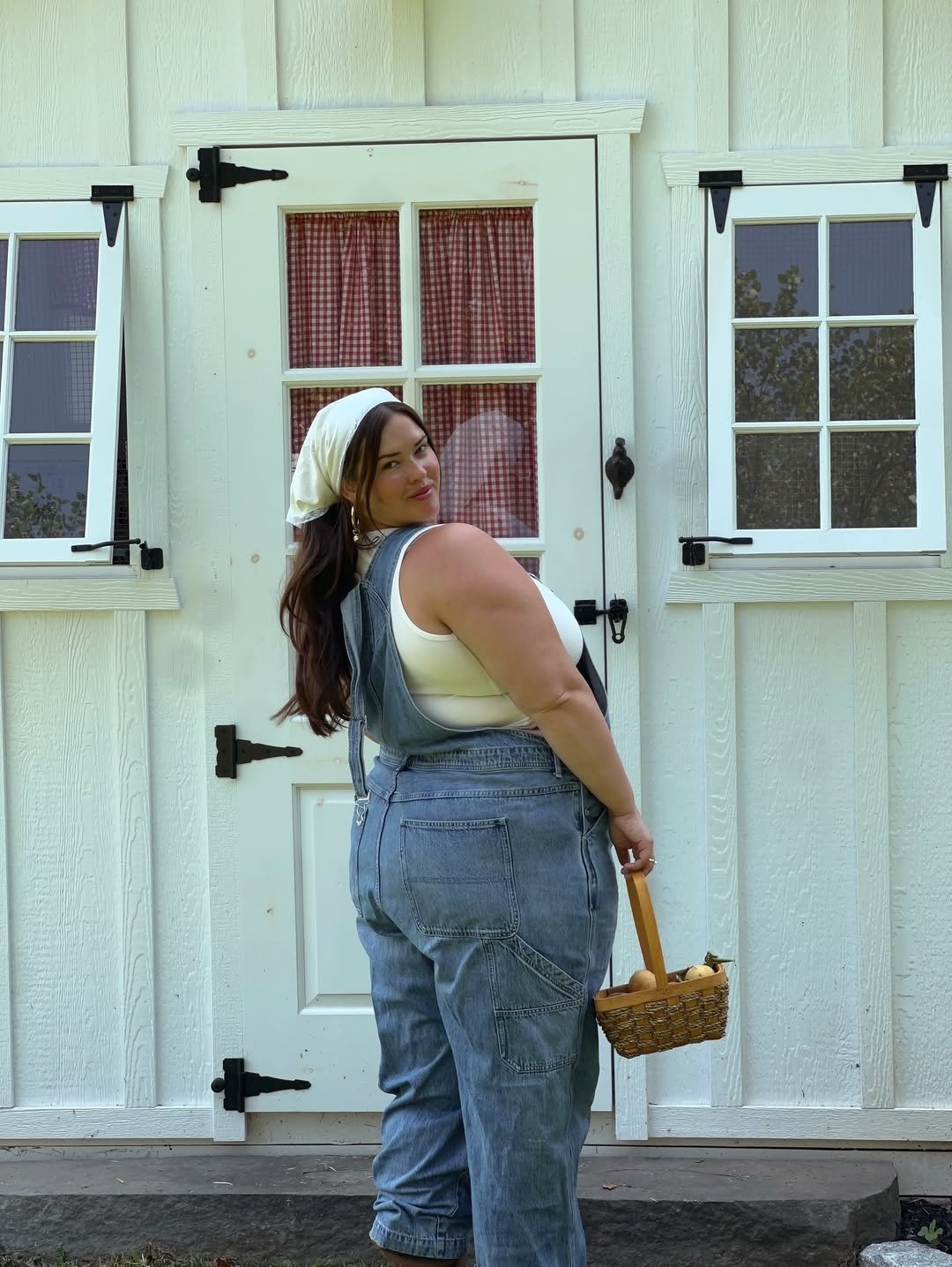 A imagem apresenta uma mulher usando um macacão estiloso de jeans, que combina conforto e elegância. O macacão, com alças ajustáveis e bolsos frontais, é ideal para um visual casual. Ela segura uma cesta, adicionando um toque rústico. O fundo com uma port