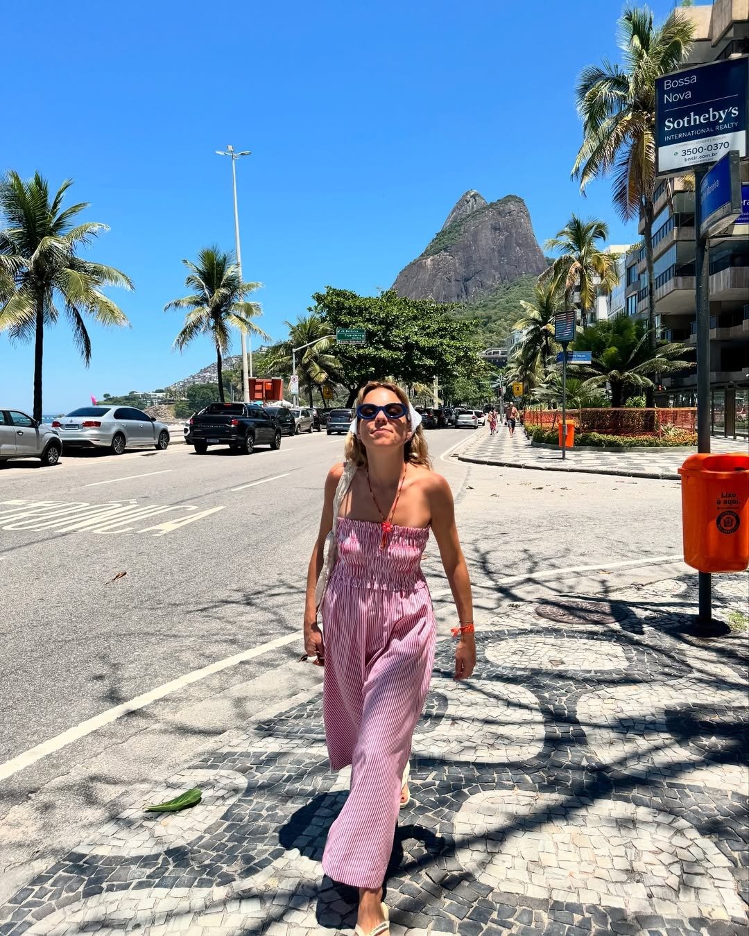 Em um cenário ensolarado, uma mulher caminha confiante em uma calçada de pedras portuguesas, vestindo um macacão estiloso listrado em rosa e branco. O modelo, leve e confortável, destaca-se pelo design moderno, ideal para dias quentes. Ao fundo, palmeiras