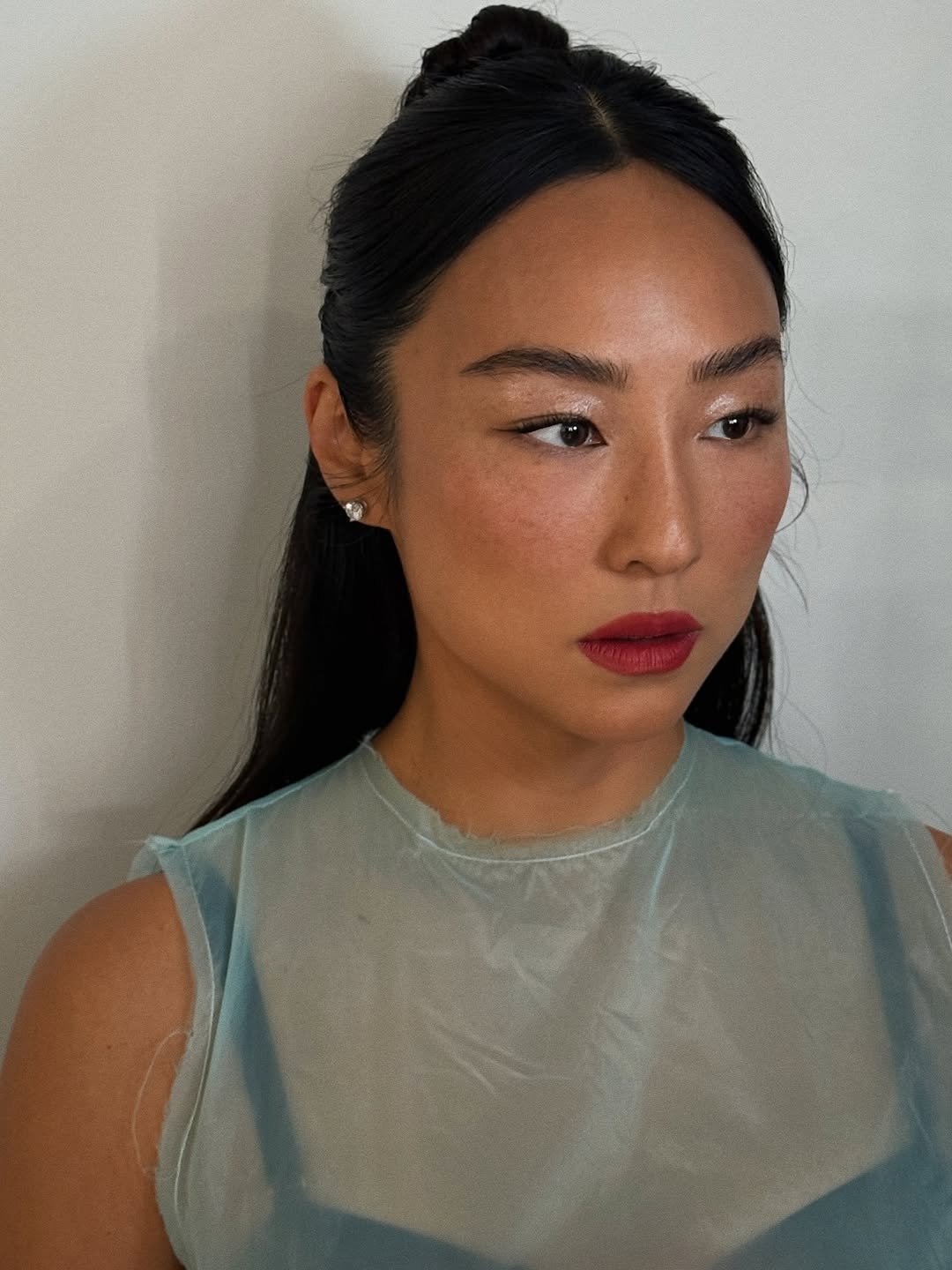 O estilo da Greta Lee se destaca pela combinação de elegância e modernidade. Nesta imagem, ela usa uma blusa de tecido transparente, que traz leveza ao visual. A maquiagem é sutil, com ênfase nos lábios em tom vermelho, contrastando com a pele luminosa. O