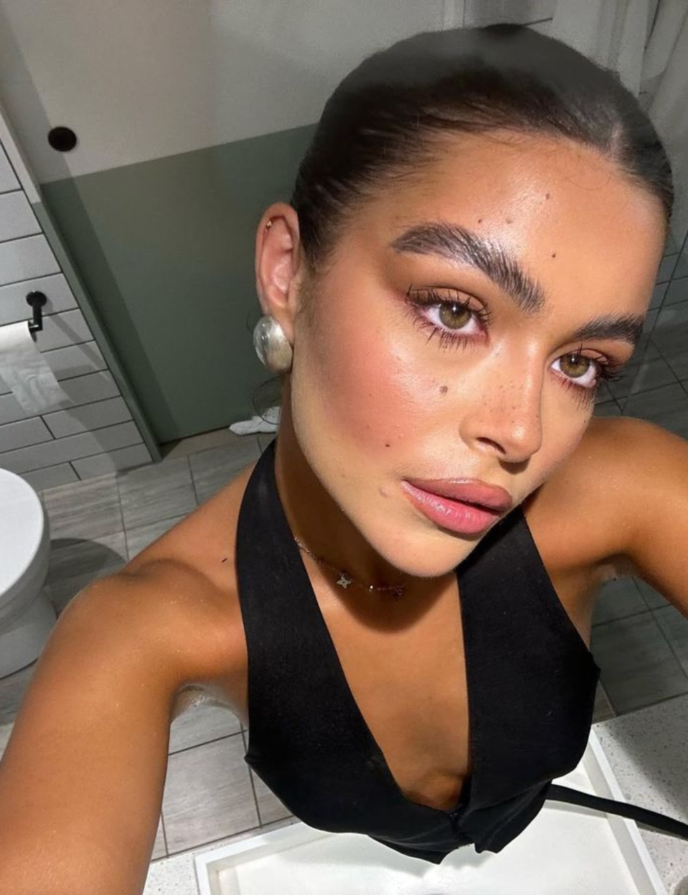 A maquiagem marrom é destaque nesta imagem, evidenciando um look sofisticado e moderno. Os olhos são realçados com sombras marrons e delineado suave, complementados por cílios volumosos. A pele tem um acabamento iluminado, com blush sutil e lábios rosa.