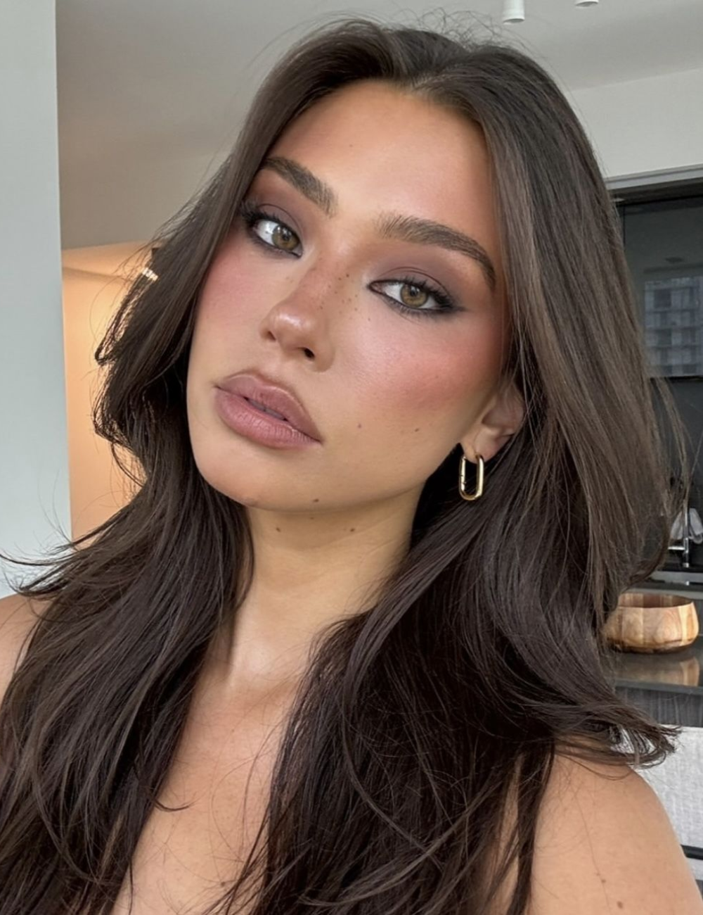 A maquiagem marrom destaca-se nesta imagem, com um olhar esfumado que realça os olhos. A pele tem um acabamento saudável, com um toque de blush rosado. Os lábios são preenchidos com um tom nude, complementando o visual.