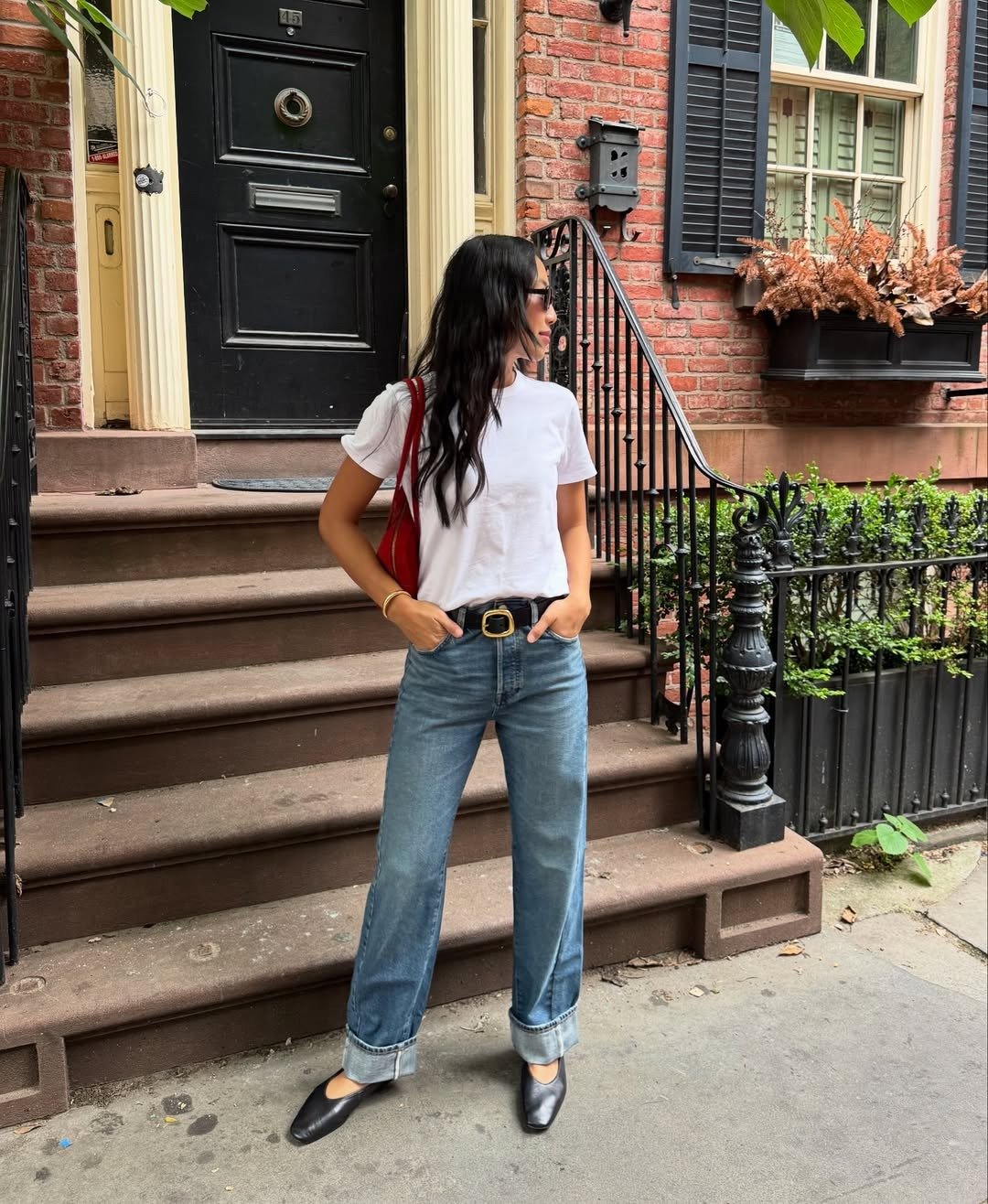 A imagem apresenta uma mulher em um ambiente urbano, usando uma camiseta branca básica e jeans de cintura alta com barras dobradas, combinados com sapatos pretos elegantes e um cabelo hidratado, resultado da máscara de cabelo coreana