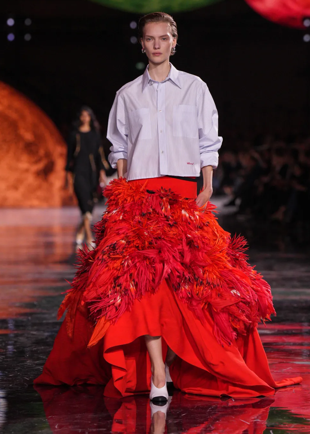 Desfile de moda com modelo em camisa larga e saia vermelha com detalhes de plumas, estilo Matthieu Blazy.