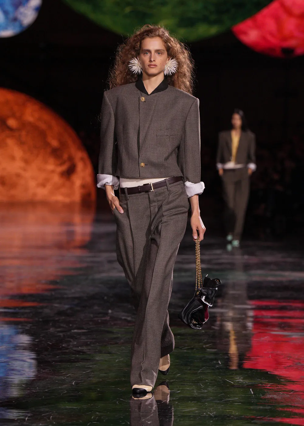 Modelo com terno cinza na passarela, bolsa preta. Cenário colorido ao fundo. Matthieu Blazy.