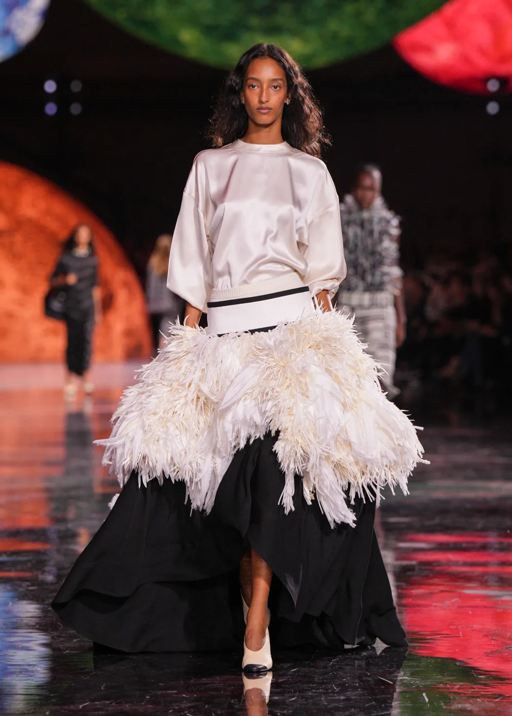 Modelo desfilando com saia volumosa com plumas brancas e blusa de cetim, em coleção de Matthieu Blazy.