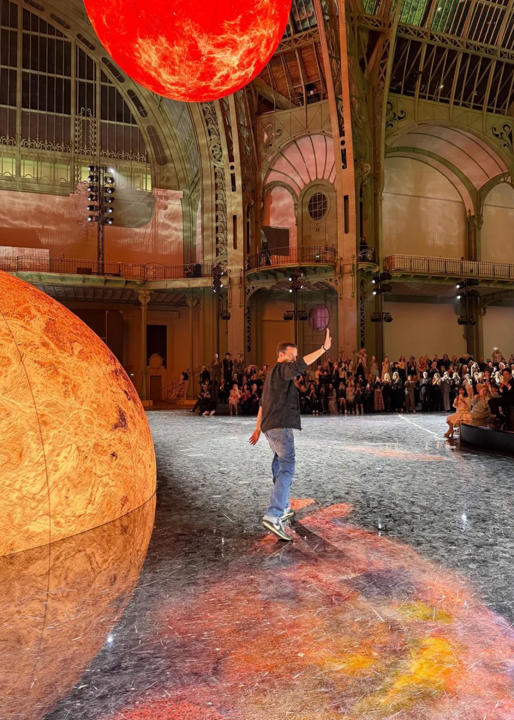 Grande salão com estrutura metálica, esferas gigantes no teto e pessoa de costas acenando ao público.