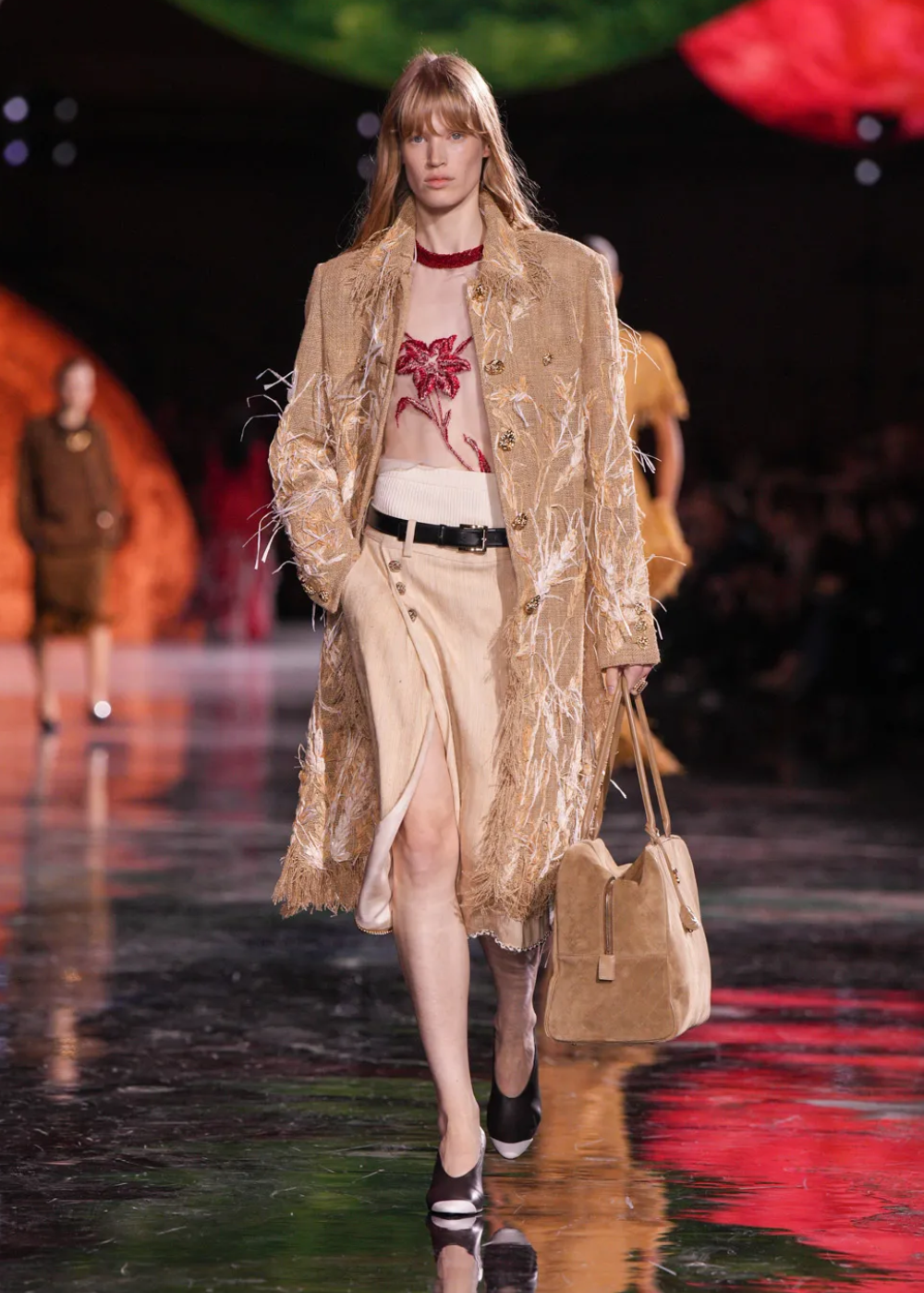 Modelo desfila em passarela usando casaco texturizado e saia bege, segurando bolsa, em evento de moda.