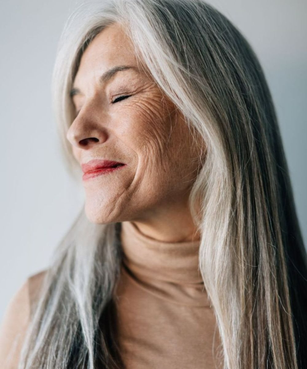 Uma mulher com cabelos longos e grisalhos, usando um suéter de gola alta em tom neutro, exibe uma expressão serena. Seus olhos estão fechados, destacando a suavidade da pele tratada com creme para área dos olhos