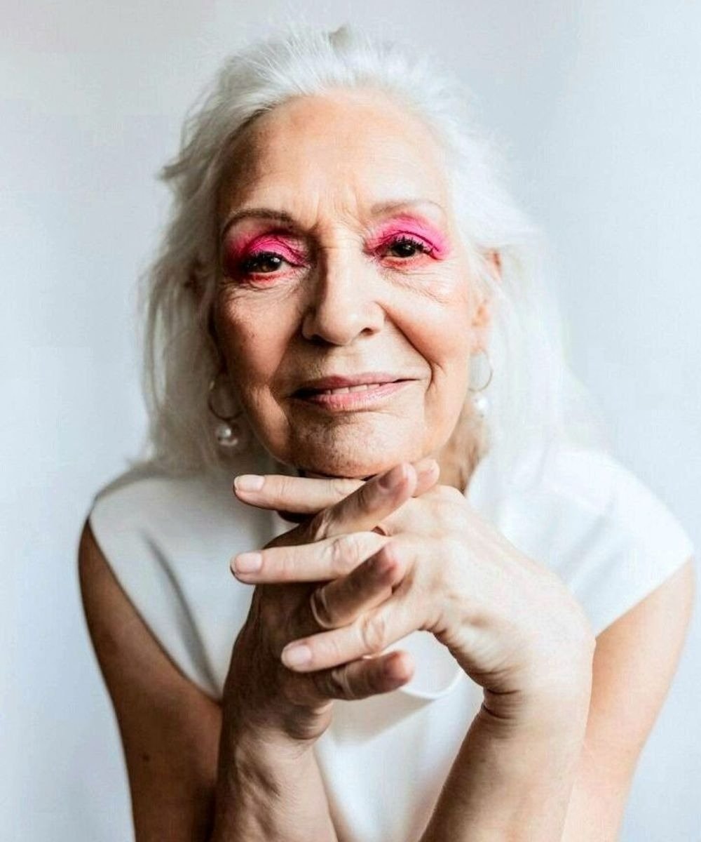 Uma mulher idosa com cabelo longo e branco exibe um olhar confiante, destacando-se com maquiagem vibrante, depois de usar creme para área dos olhos
