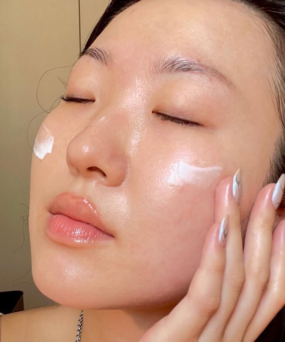 A imagem mostra uma mulher aplicando protetor solar no rosto, destacando a importância dos cuidados com a pele.