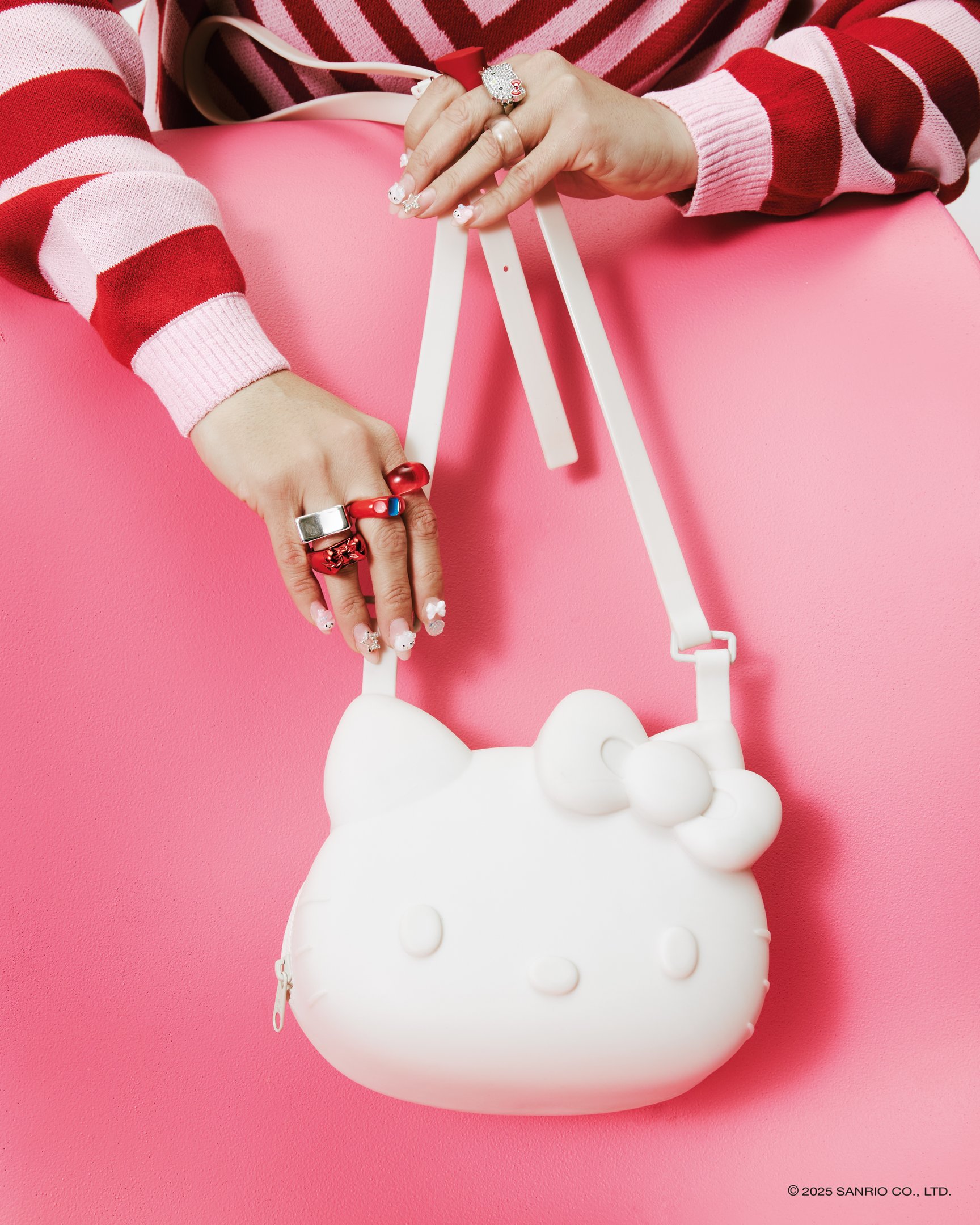 Bolsa Hello Kitty branca com detalhes Melissa, em um fundo rosa. Unhas decoradas e anéis coloridos adornam as mãos.
collab Melissa e Hello Kitty