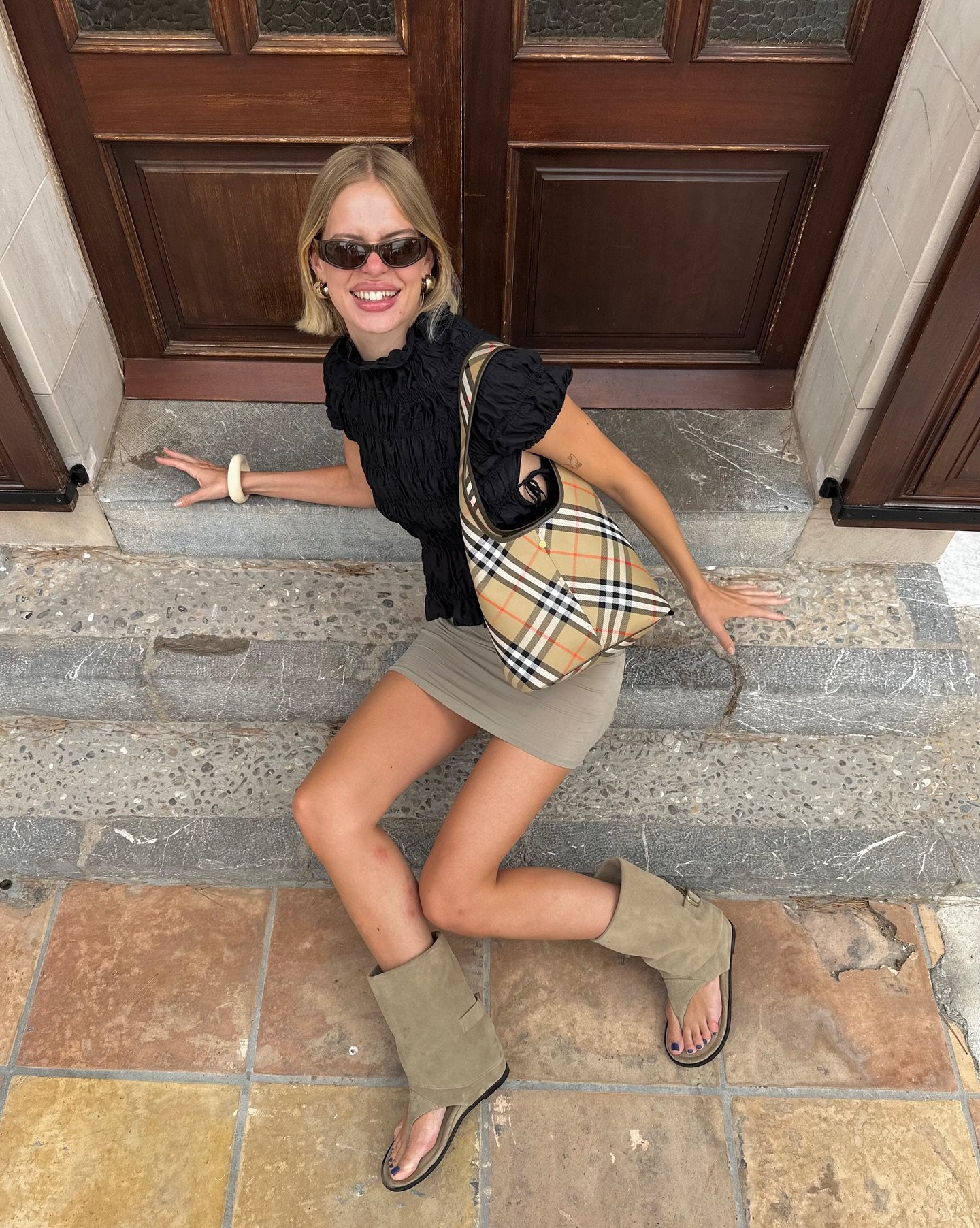 Pessoa sorrindo sentada em degraus, usando bolsa xadrez e óculos de sol. As botas de estilo Miu Miu completam o look.