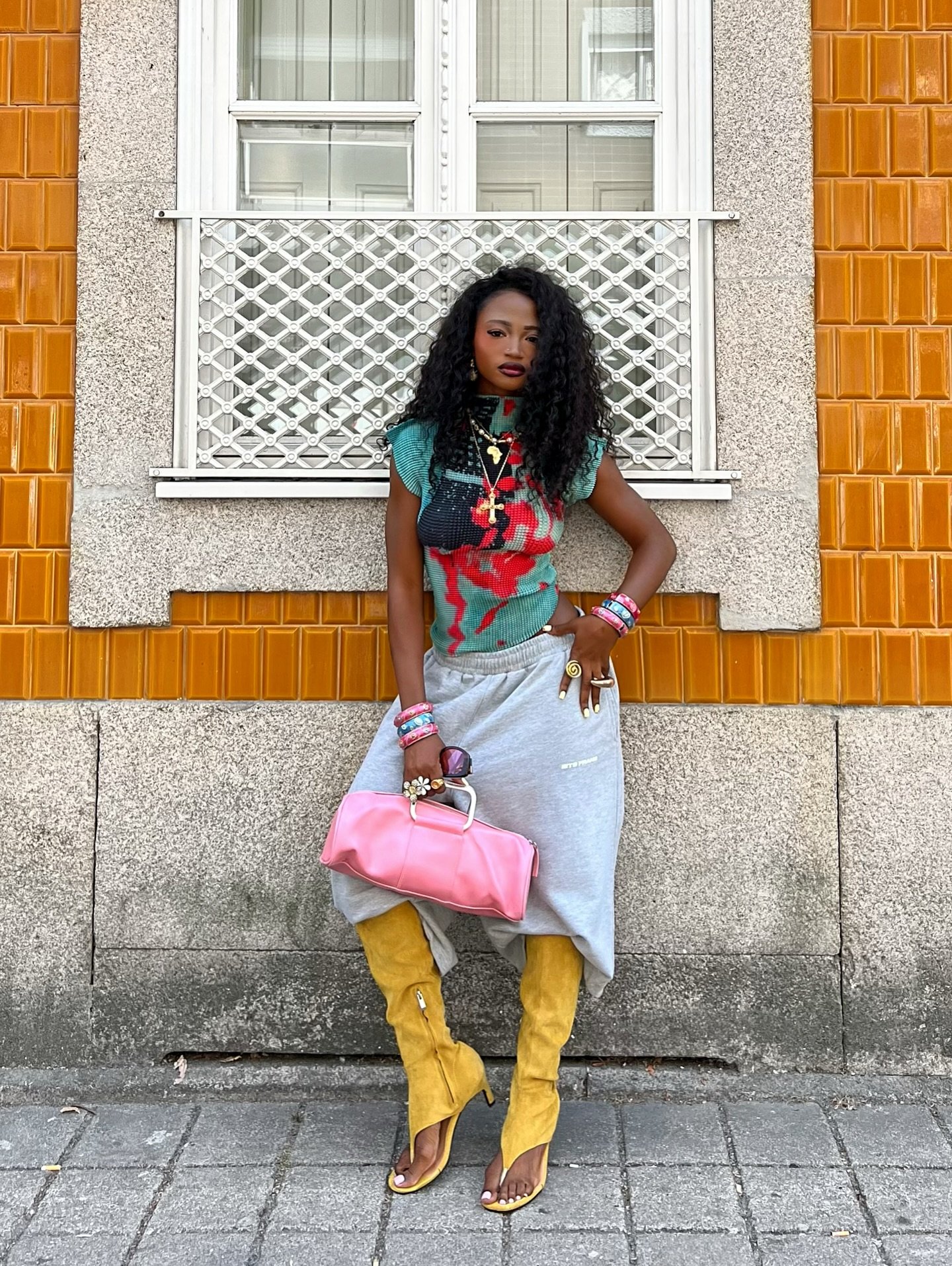 Mulher com cabelo cacheado veste blusa estampada e saia cinza, segura bolsa rosa, e usa botas amarelas. Miu Miu ausente.