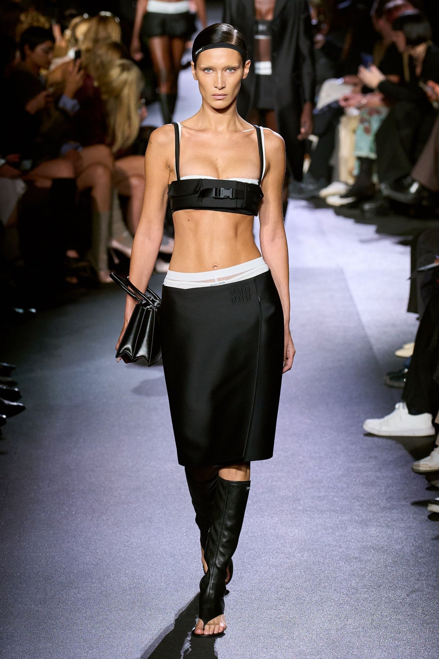 Modelo desfila em um evento de moda usando conjunto preto Miu Miu, com saia e top, acompanhados de botas de cano alto.