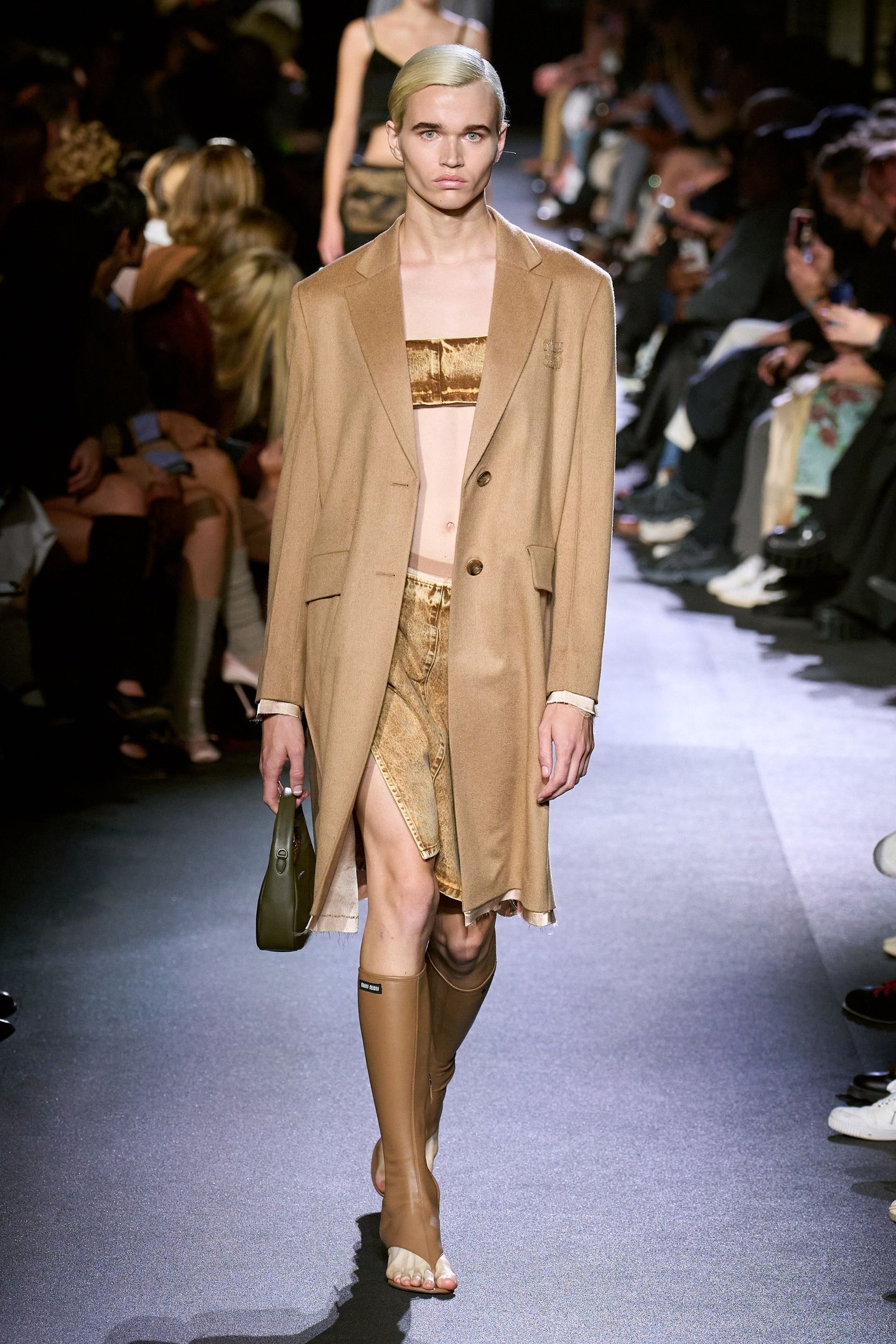 Desfile de moda com modelo usando casaco bege, conjunto dourado e sandálias marrons.