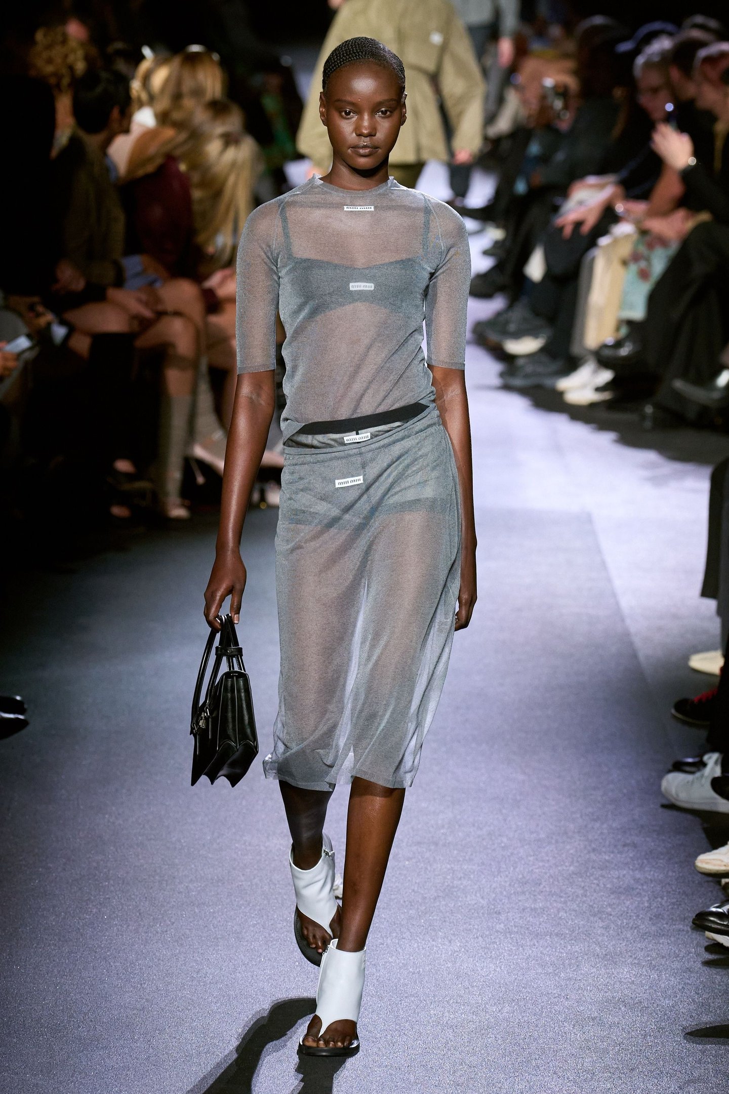Modelo desfila usando vestido transparente azul, carregando bolsa preta. Coleção Miu Miu em destaque.