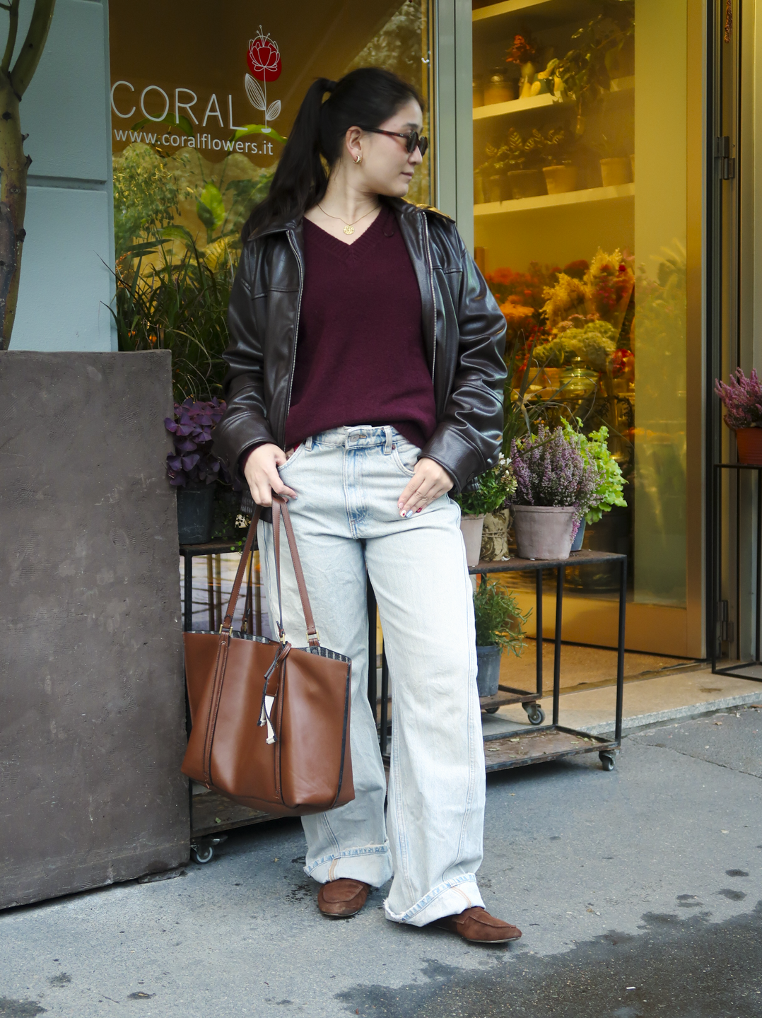 Jovem em frente a floricultura usando jaqueta marrom, blusa vinho e jeans. Cenário lembra Milão Fashion Week.