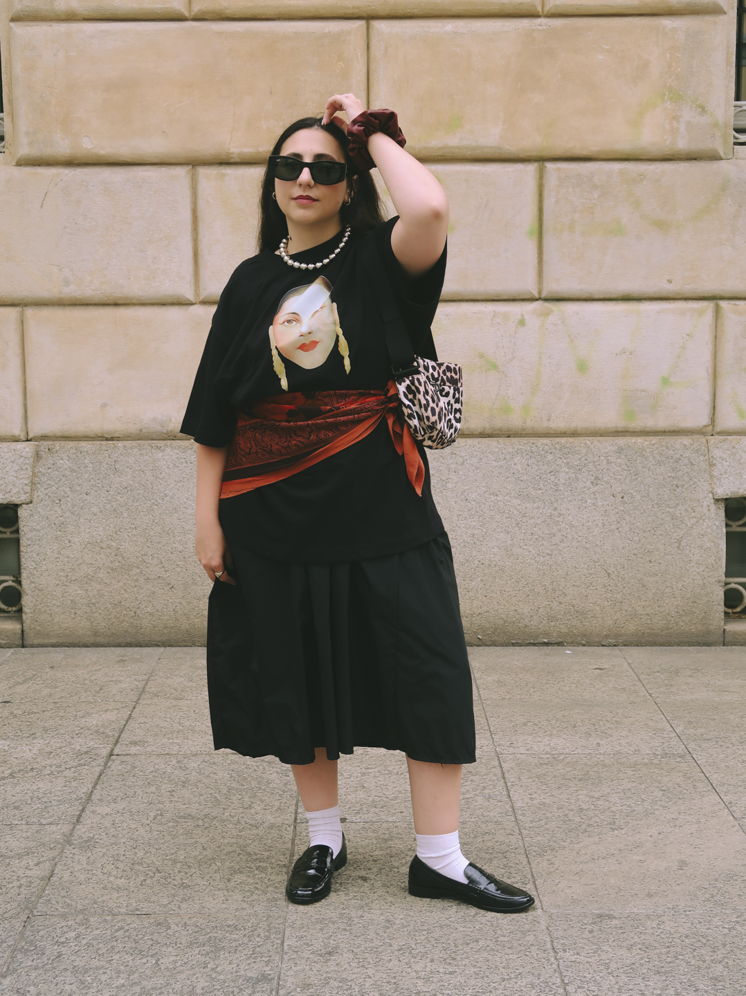 Pessoa com look estiloso, usando camiseta gráfica, saia midi preta e óculos escuros. Inspiração Milão Fashion Week.