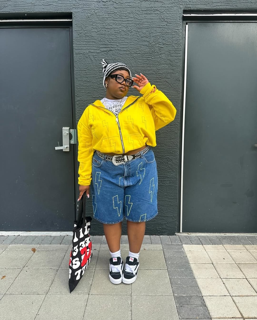 A moda anos 2010 é representada por um look vibrante e ousado. A pessoa usa um moletom amarelo com detalhes em relâmpago, combinado com uma saia jeans oversized, também decorada com o mesmo tema.