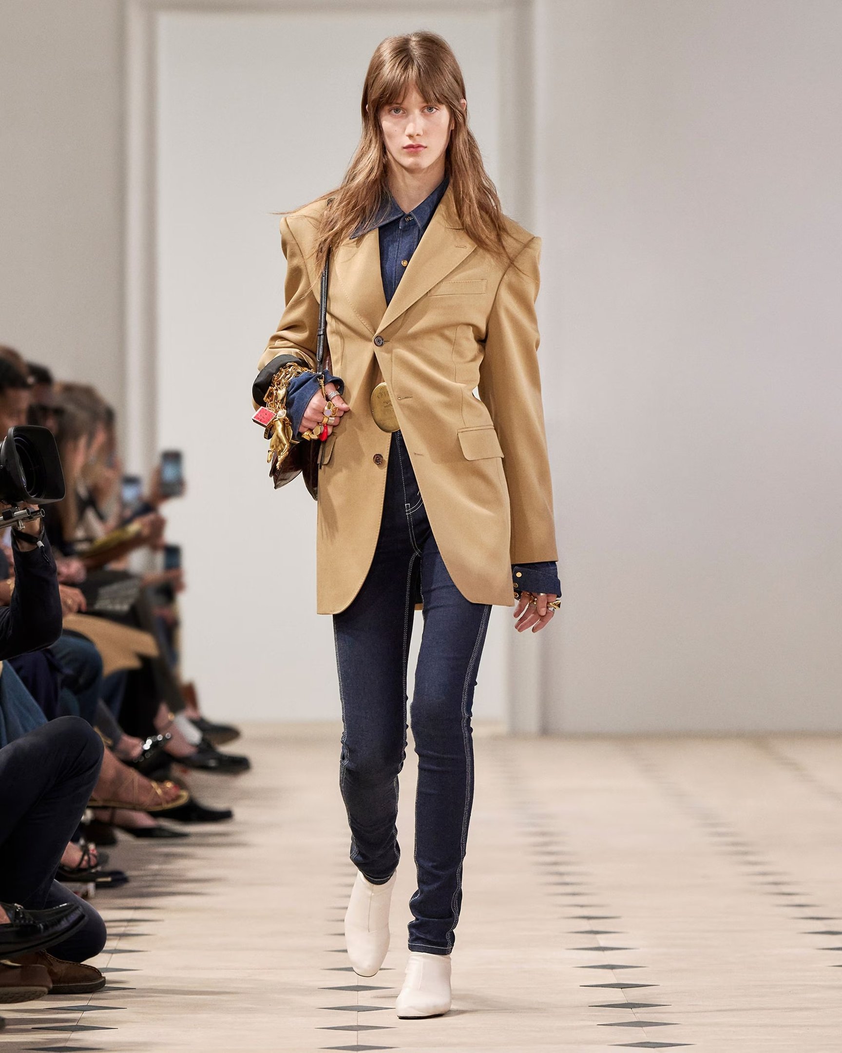 A moda anos 2010 se destaca pela mistura de estilos e pela ousadia nas combinações. Nesta imagem, uma modelo desfila usando um blazer oversized em tom bege, que remete à estética retrô, combinando com uma camisa jeans e calças skinny escuras.