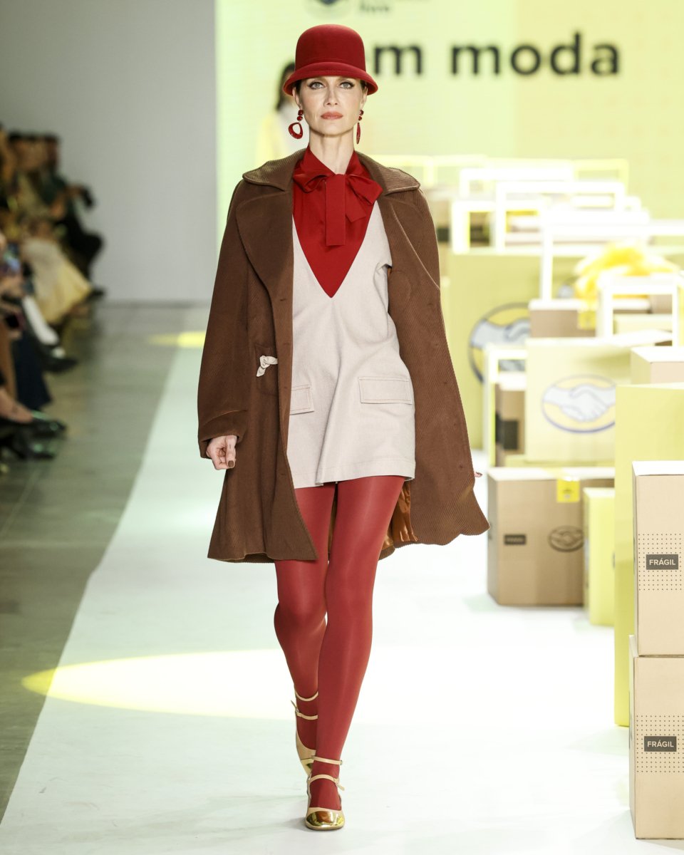 Modelo desfila em passarela com um visual que remete à elegância vintage história da moda brasileira. Ela usa um vestido claro com um laço vermelho, acompanhado de um casaco marrom e meia-calça vermelha. O chapéu vermelho e brincos grandes completam o look, que ev