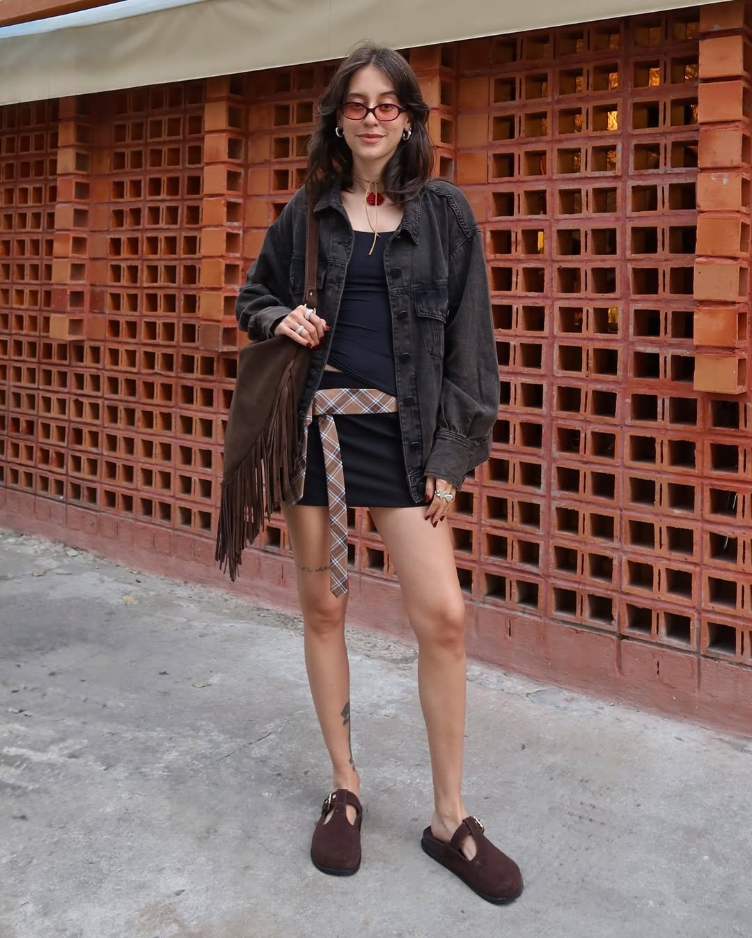 Mulher com look com referências da moda anos 60, com óculos pequenos, bolsa de franjas e colar maxi em destaque.