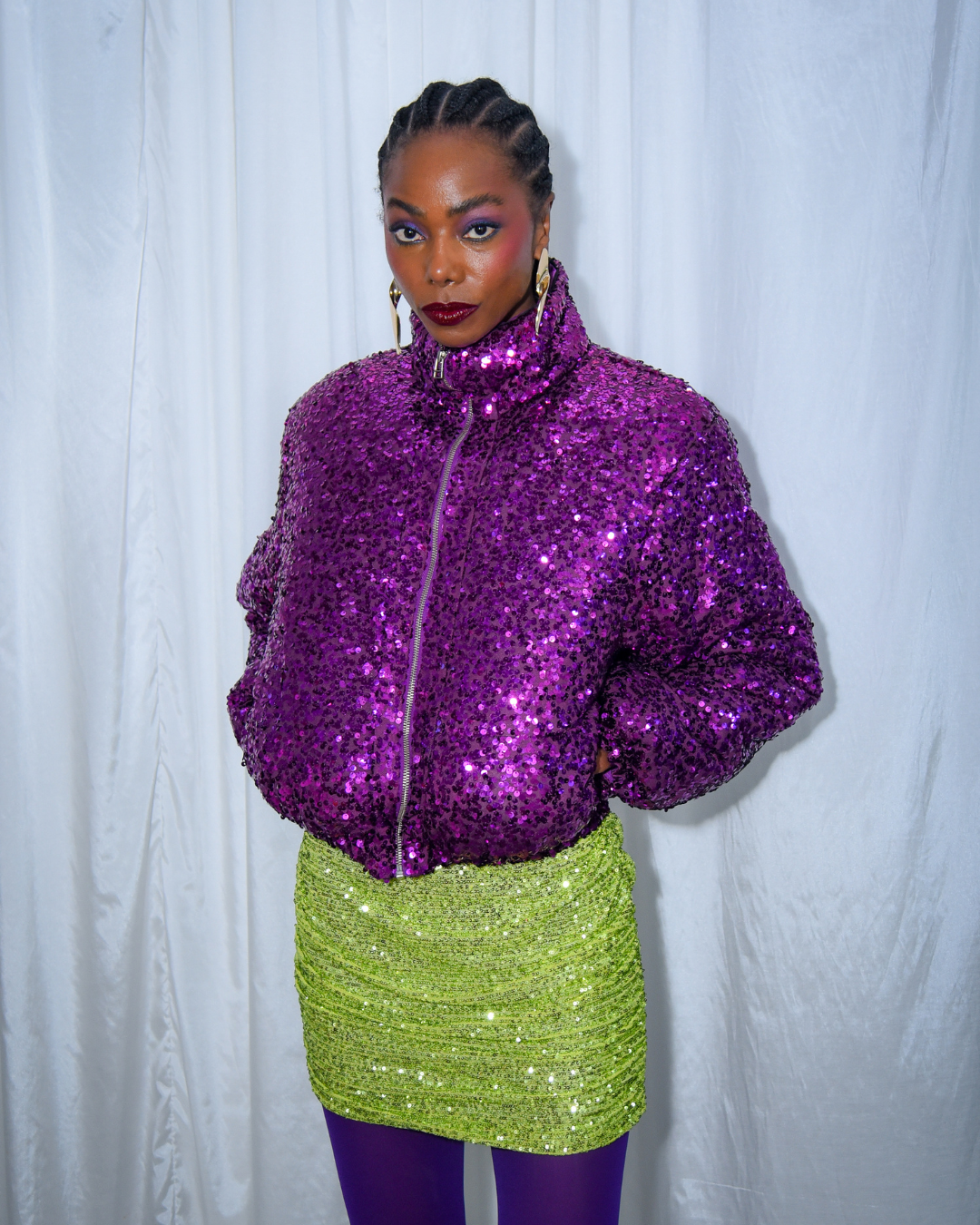 A imagem apresenta uma modelo vestindo um casaco roxo brilhante, coberto de paetês, e uma saia verde neon, também com detalhes cintilantes. A combinação vibrante e ousada reflete a influência da cultura pop e da moda contemporânea da história da moda brasileira