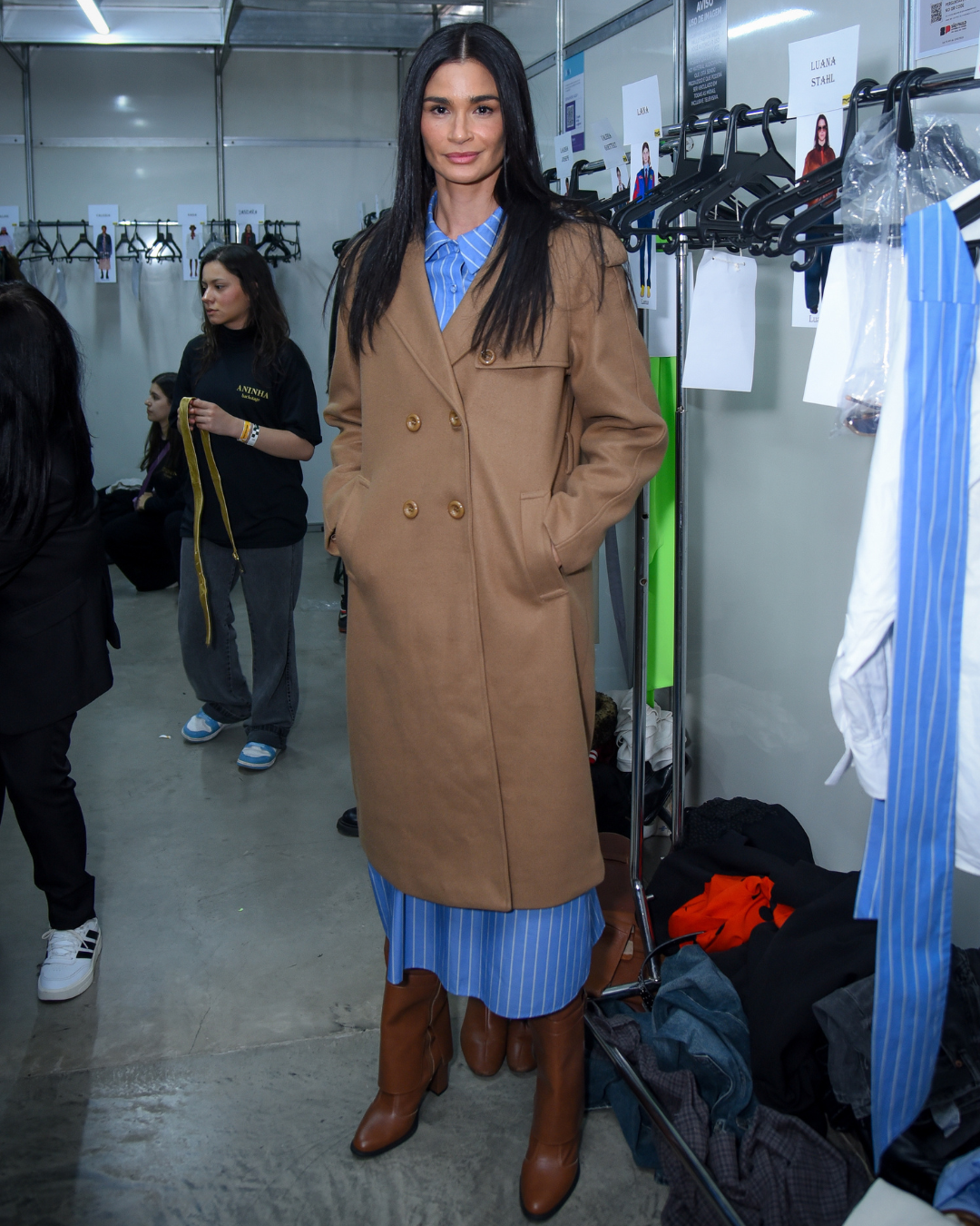 A imagem retrata uma modelo em um ambiente de backstage de moda, destacando a fusão de estilos contemporâneos na história da moda brasileira. Ela usa um casaco longo e clássico na cor caramelo, combinado com uma camisa listrada azul, que reflete a tendênc