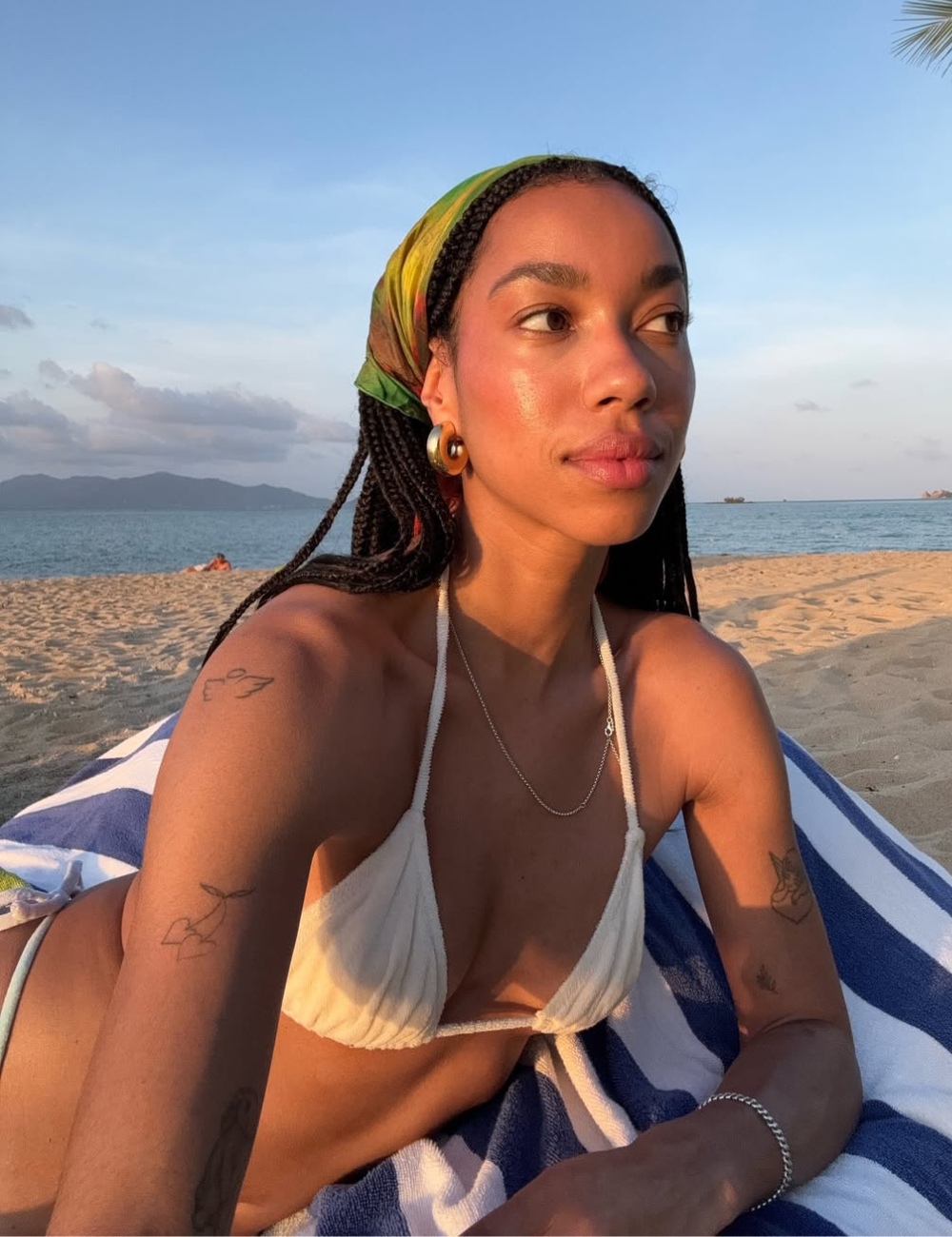 A imagem apresenta uma jovem com cabelo trançado, usando um biquíni branco e um lenço colorido na cabeça, em um ambiente de praia ao pôr do sol. O cenário destaca a beleza da moda praia 2026, com ênfase em peças minimalistas e acessórios elegantes.