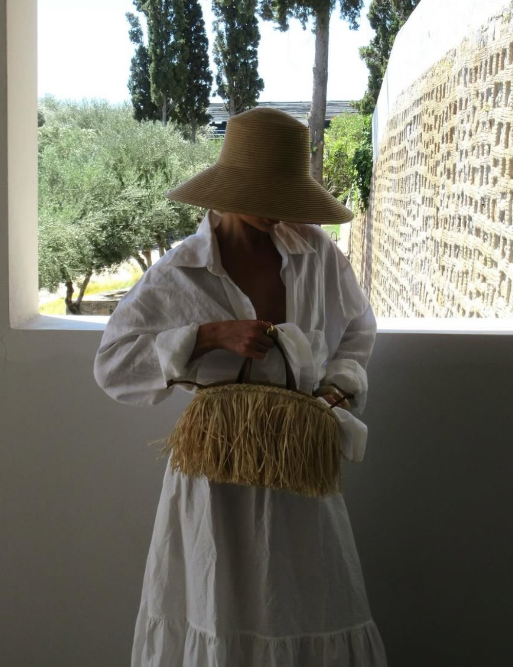 A imagem retrata uma mulher vestindo uma camisa branca oversized, ideal para a moda praia 2026. O destaque vai para o chapéu de palha grande, que oferece proteção solar e estilo. Ela segura uma bolsa de palha trançada.