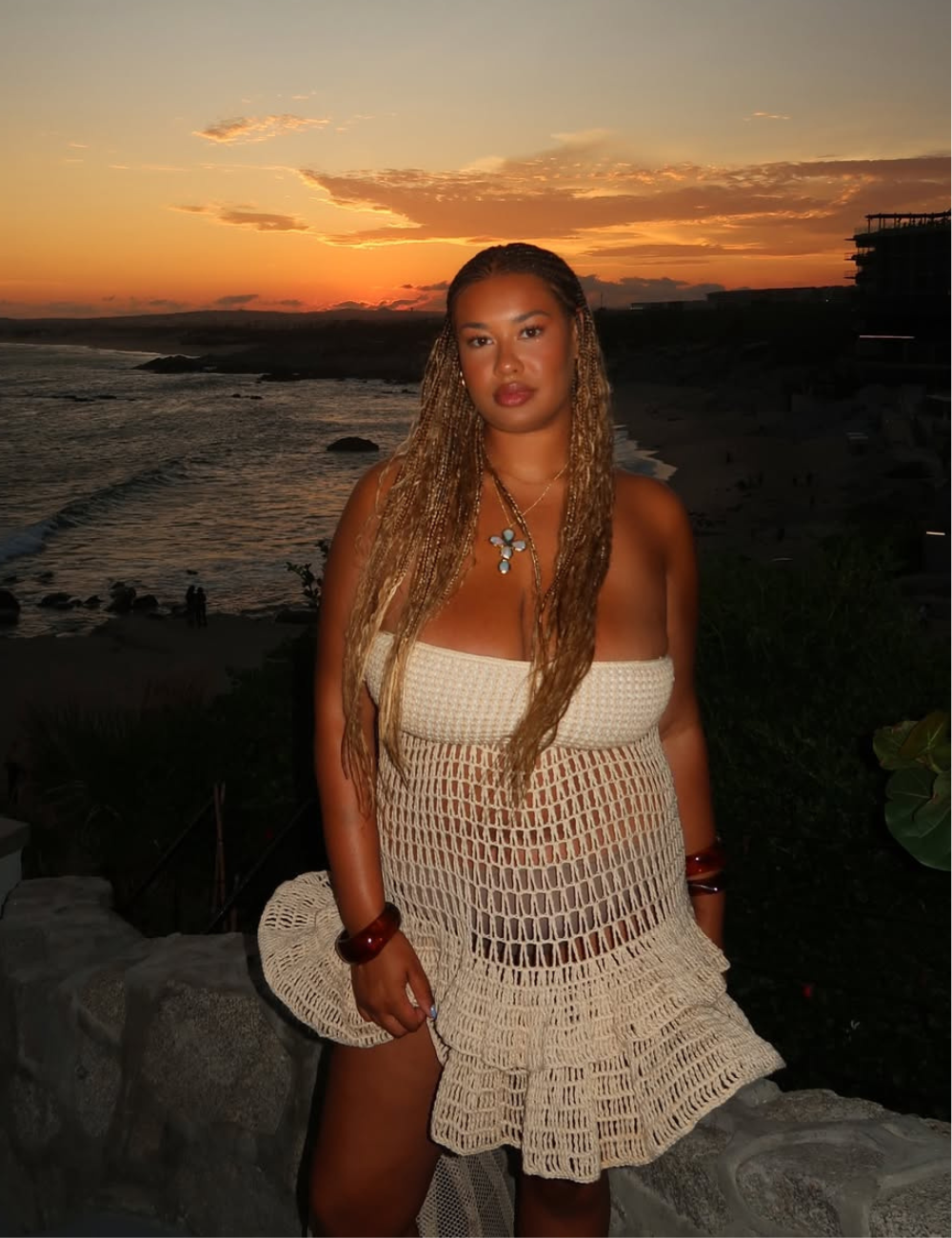 A imagem destaca uma modelo em um cenário de pôr do sol na praia, vestindo um vestido de crochê na cor bege, que reflete as tendências de moda praia 2026. O vestido, com detalhes vazados e uma saia rodada, combina conforto e estilo.