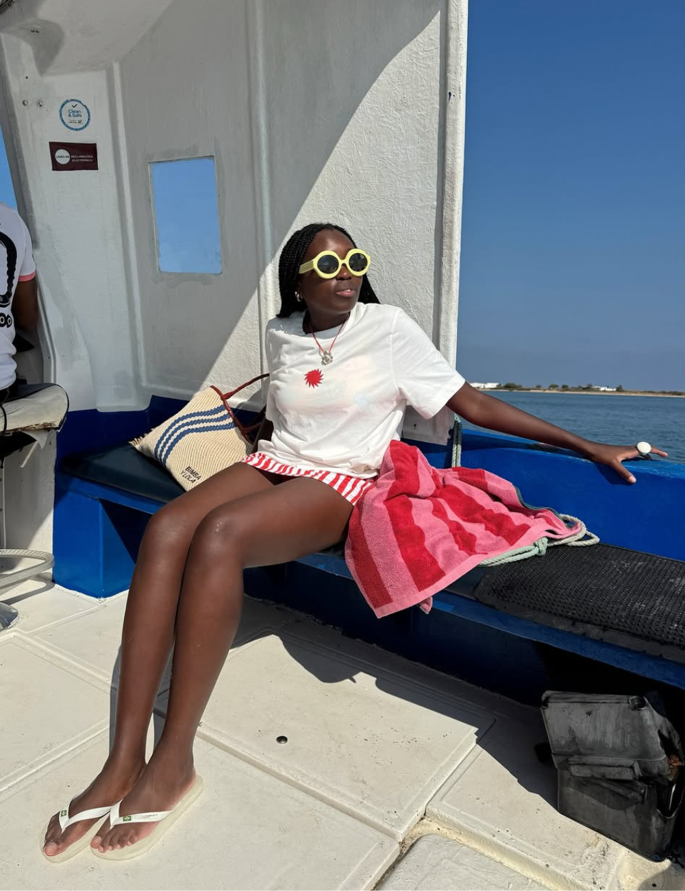 A moda praia 2026 destaca-se pela combinação de conforto e estilo. Na imagem, uma mulher usa uma camiseta branca com estampa de sol, shorts listrados em vermelho e branco, e óculos de sol grandes, trazendo um ar descontraído e moderno.