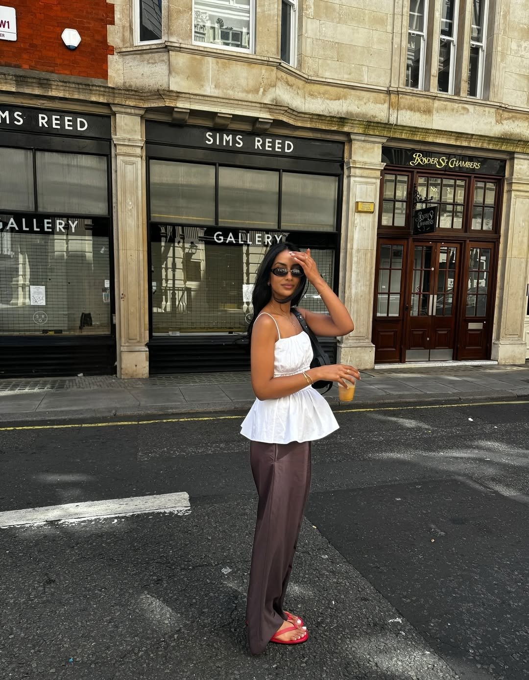 Uma jovem modelo exibe uma blusa branca de tecido leve, com detalhes peplum, ideal para o verão. Acompanhada de calças largas em tom marrom e sandálias vermelhas, ela complementa o look com óculos escuros e uma bolsa preta. Modelos de blusa para verão