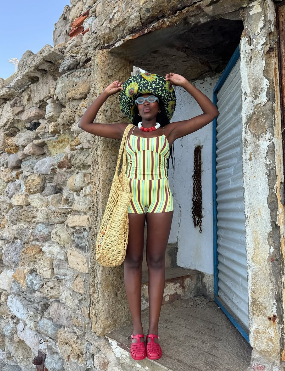 Modelos de bolsa de crochê para praia são tendência nesta imagem, onde uma mulher exibe uma bolsa de crochê em tom natural, ideal para compor looks de verão. Ela veste um maiô listrado vibrante e um chapéu grande com estampa tropical.