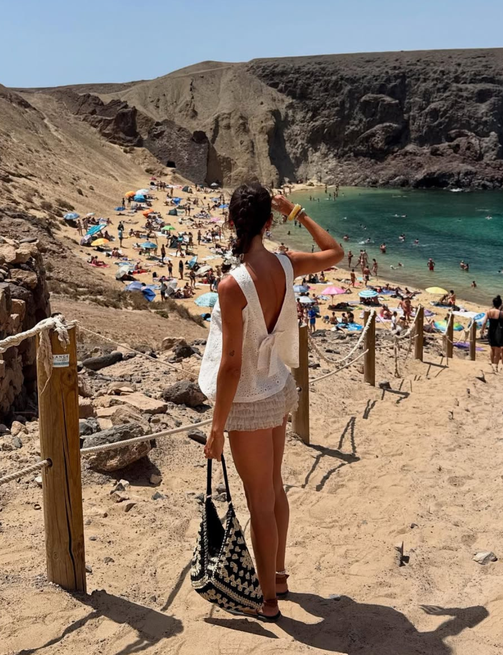 Uma mulher está de costas, usando um top branco de crochê e shorts claros, enquanto desce uma trilha de areia em direção à praia. Ela segura uma bolsa de crochê em preto e branco, que complementa seu visual casual e elegante.