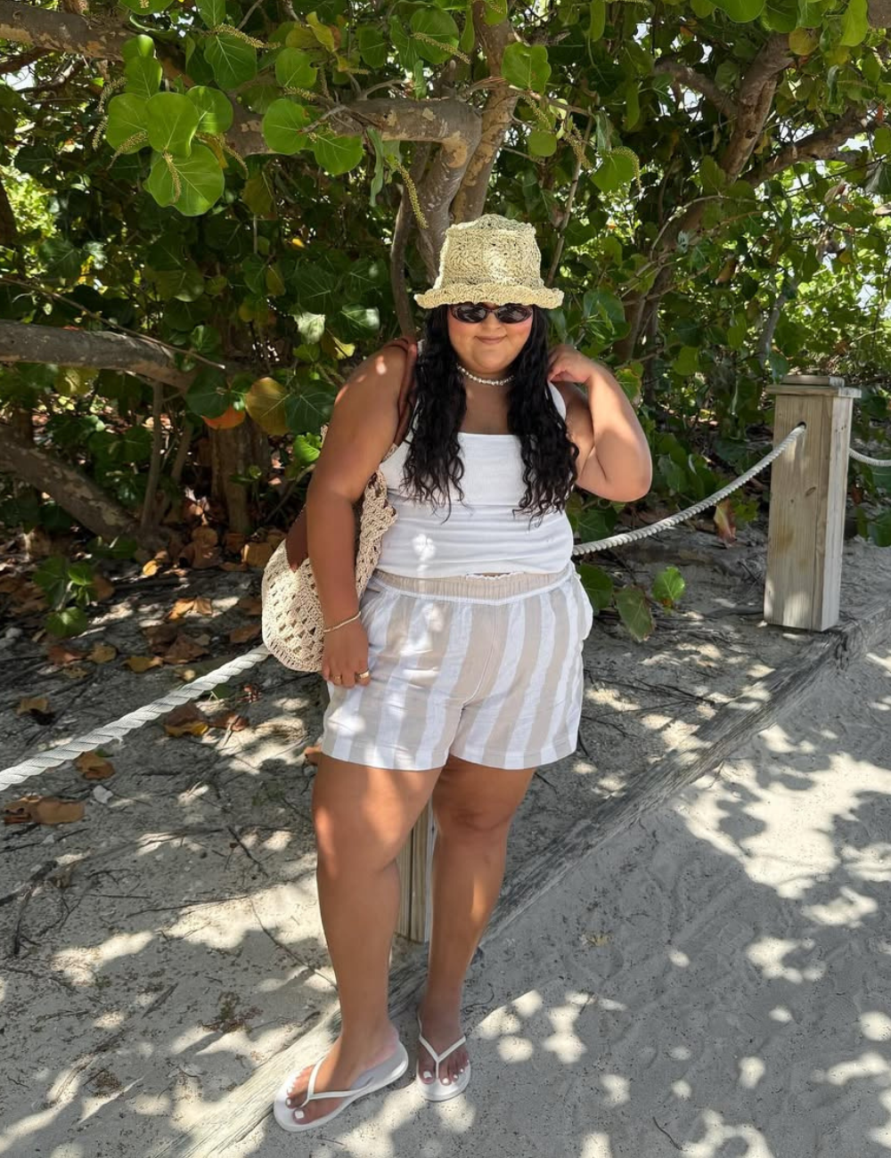A imagem apresenta uma mulher vestindo um top branco e shorts listrados em tons claros, ideal para um look de praia. Ela usa um chapéu de crochê e óculos escuros, complementando o visual. Na mão, carrega uma bolsa de crochê, perfeita para a praia. 
