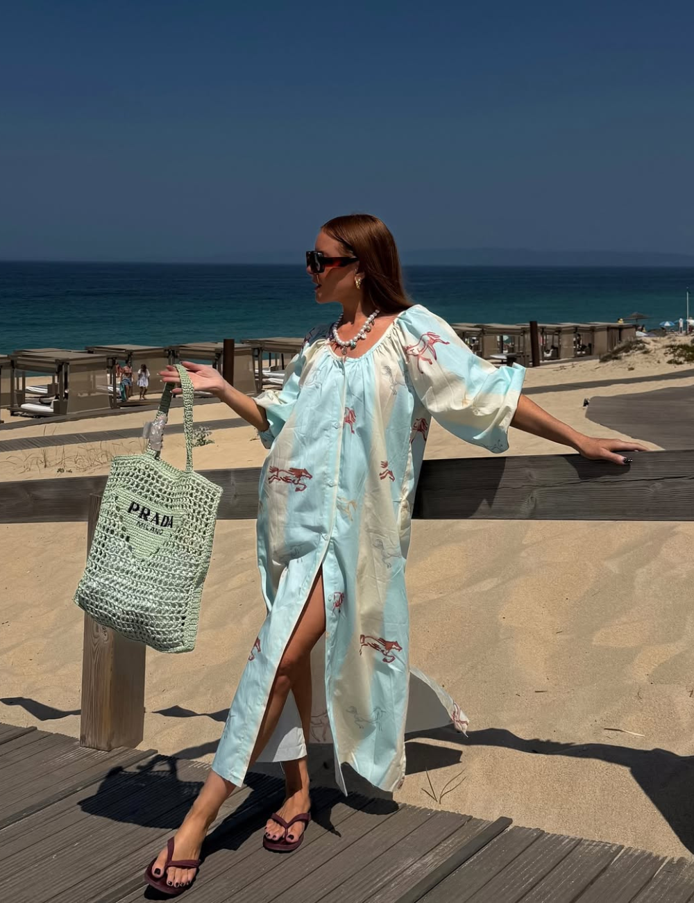 Uma mulher em uma praia deslumbrante exibe um vestido leve de cor azul claro, com detalhes florais, perfeito para o clima tropical. Ela segura um dos modelos de bolsa de crochê para praia.