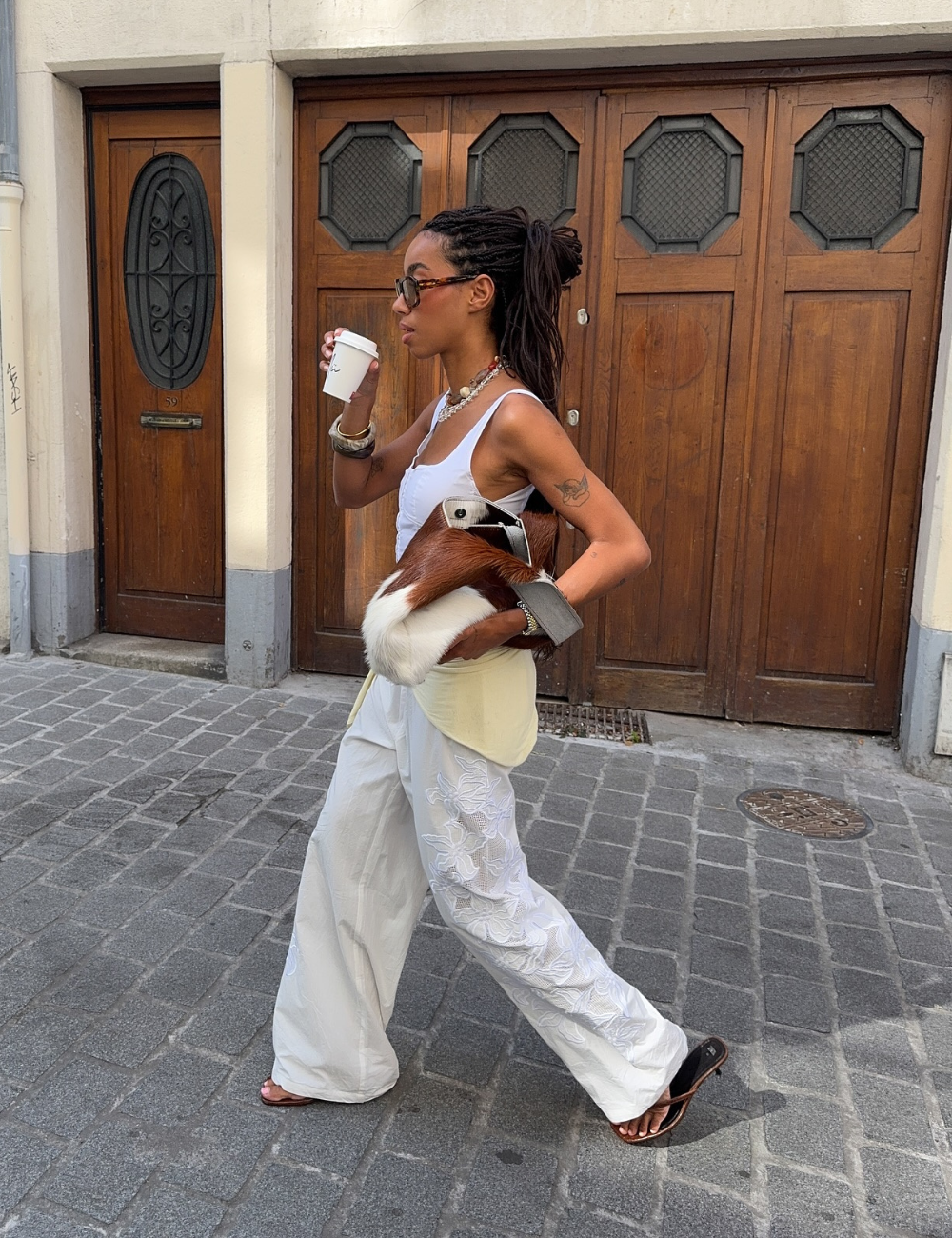 Pessoa caminha em rua de paralelepípedo usando bolsa moderna e estilosa, modelo tendência, enquanto segura um café.
