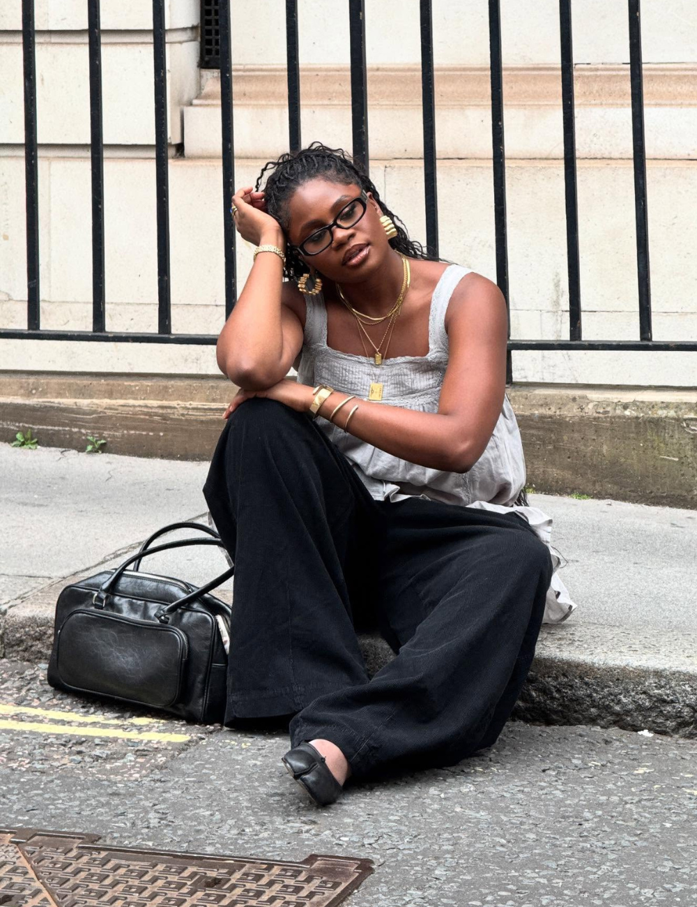 Mulher sentada na calçada com bolsa preta ao lado, usando acessórios dourados e roupas estilosas.
