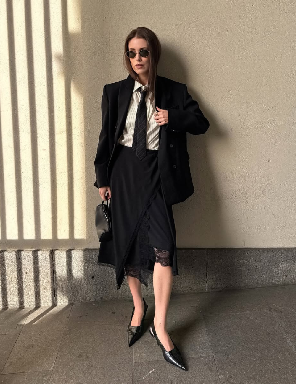 A imagem apresenta uma mulher vestindo um conjunto sofisticado, destacando modelos de saia da moda. Ela usa uma saia preta com detalhes em renda, que confere um toque elegante e moderno. O look é complementado por uma camisa listrada e um blazer oversized