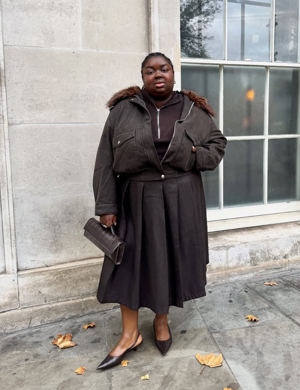 A imagem apresenta uma mulher estilosa usando uma saia midi em tom escuro, que é uma das tendências em modelos de saia da moda. Ela combina a saia com uma jaqueta oversized, complementada por detalhes em pelagem, e um suéter preto.