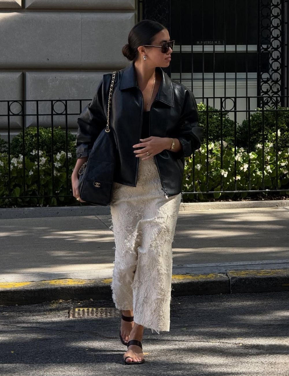 A imagem apresenta uma mulher estilosa usando uma saia longa e fluida em tom claro, com detalhes texturizados que adicionam sofisticação. Ela combina a saia com uma jaqueta de couro preta oversized, criando um contraste moderno.