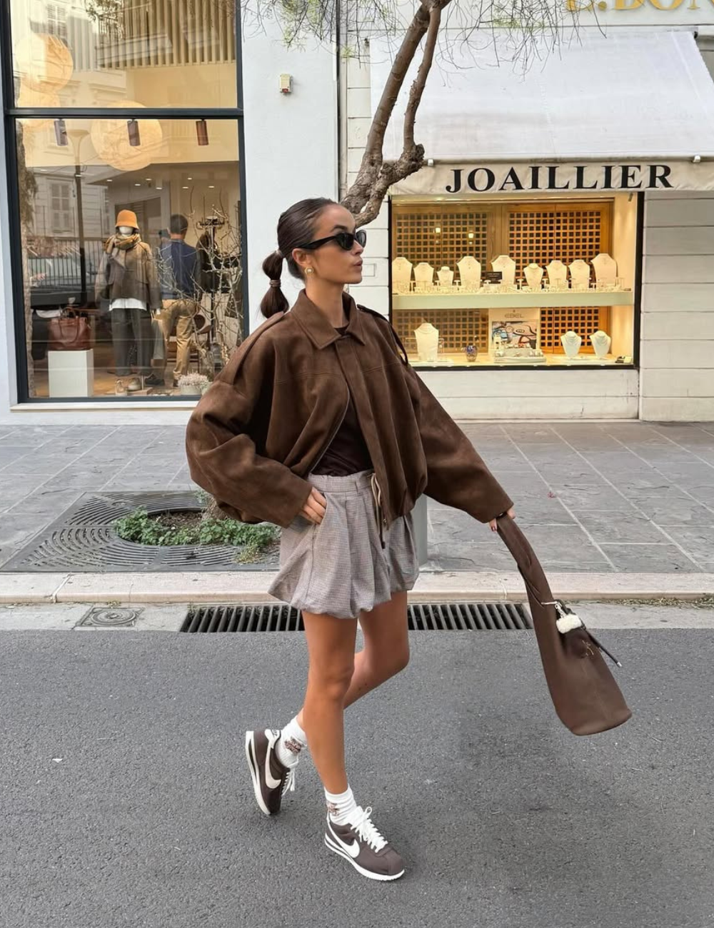 A imagem apresenta uma mulher estilosa usando um conjunto moderno que destaca os modelos de saia da moda. Ela veste uma saia curta e plissada em tons neutros, combinada com um oversized jacket marrom, criando um contraste interessante.