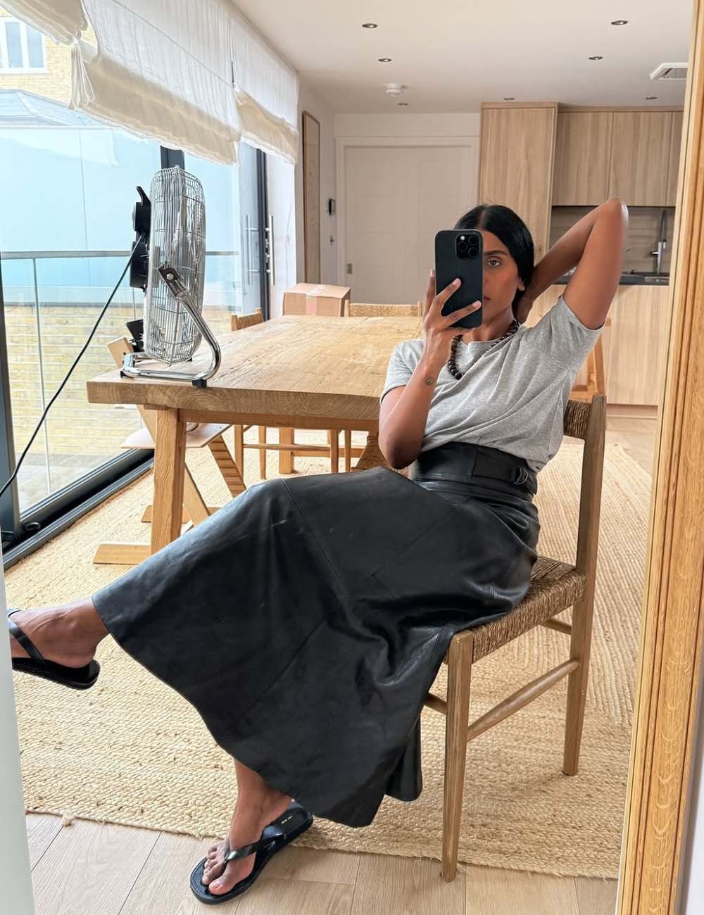 A imagem apresenta uma mulher sentada em uma cadeira de madeira, vestindo uma saia longa preta de couro e uma camiseta cinza. O look é complementado por chinelos pretos, criando um estilo casual e moderno.