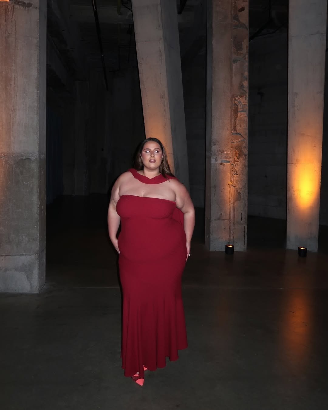 Vestido longo vermelho em ambiente industrial iluminado por velas, ressaltando um look Natal 2025.